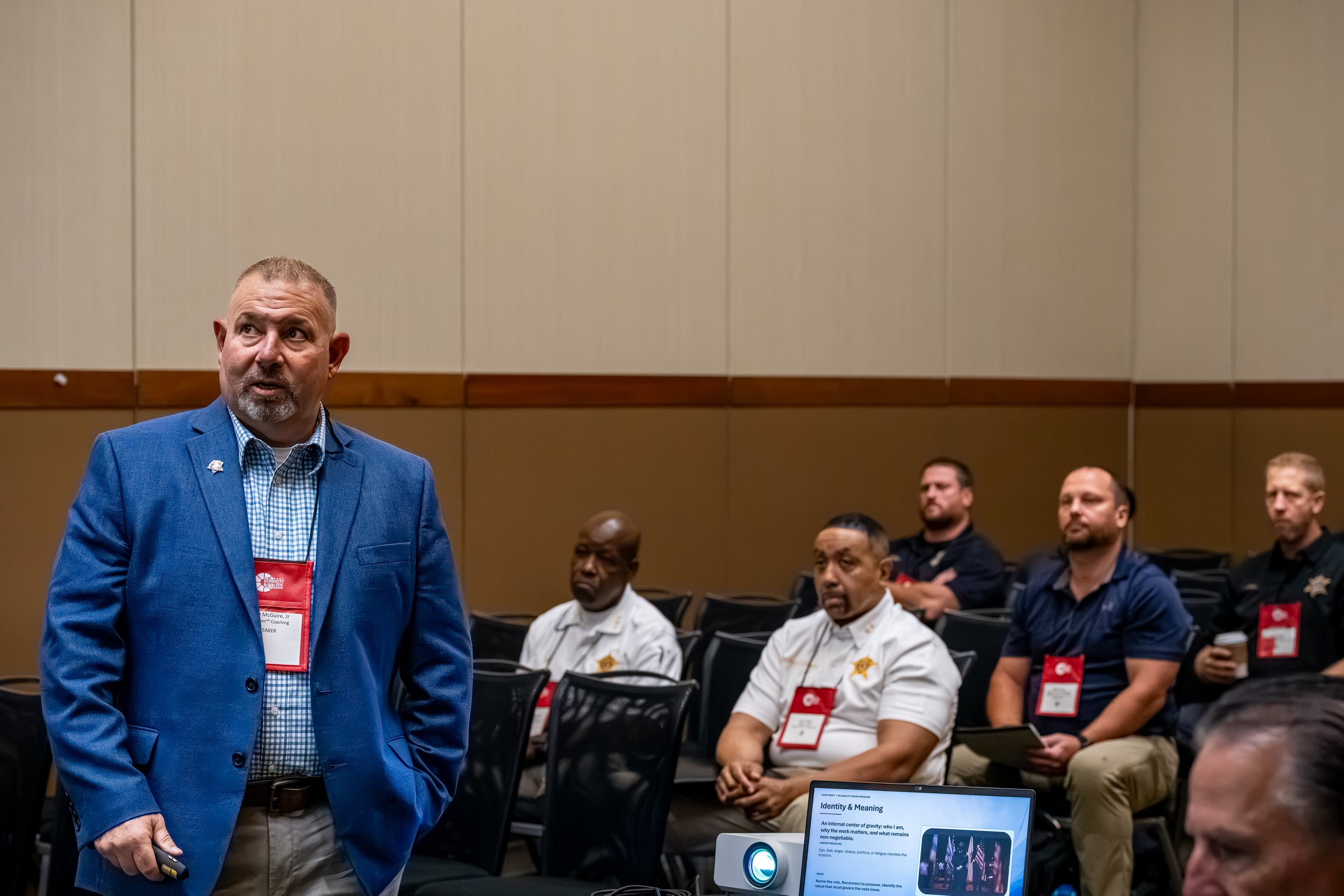 A man in a blue blazer and checkered shirt stands in front of seated people in what appears to be a conference or seminar. The background shows beige walls and several attendees wearing badges, some of whom are focused on a presentation or screen.