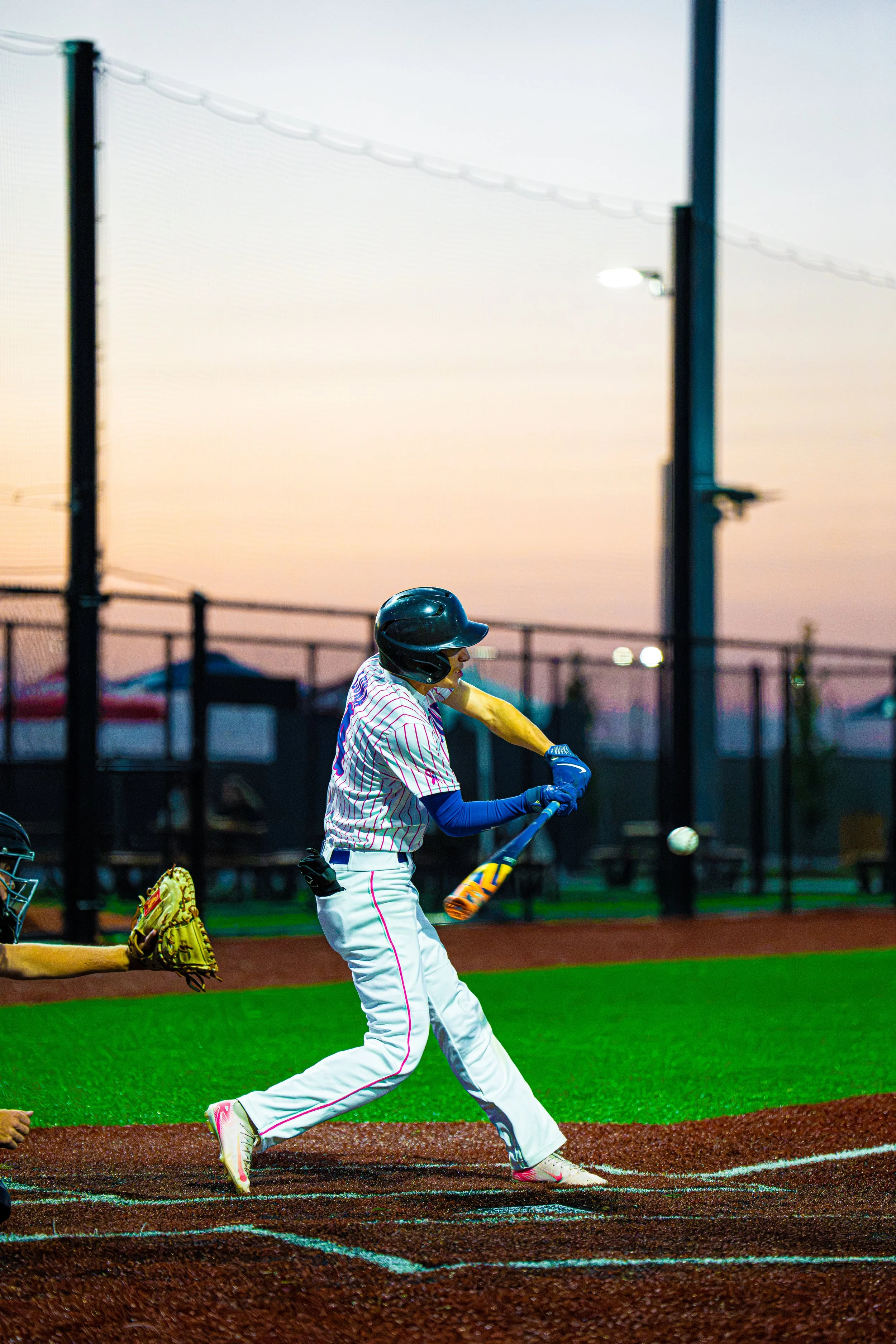 A baseball player in a striped uniform and helmet is batting on a baseball field during sunset. The batter is swinging at a pitched ball, with a catcher in a yellow mitt behind them. The background features a chain-link fence and a sunset sky.