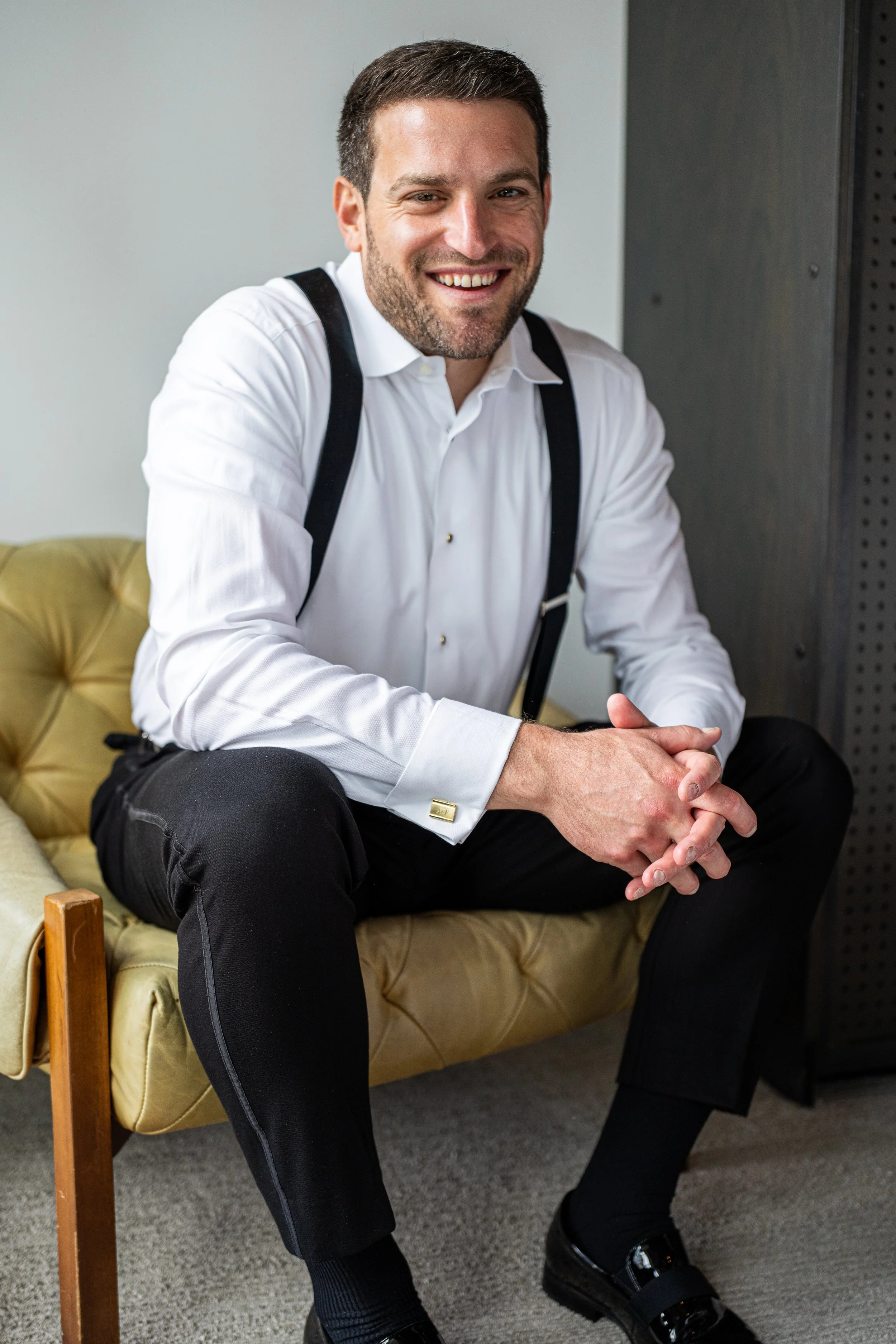 Smiling man in white shirt with black suspenders sitting on a yellow tufted chair, hands clasped, in an office setting.