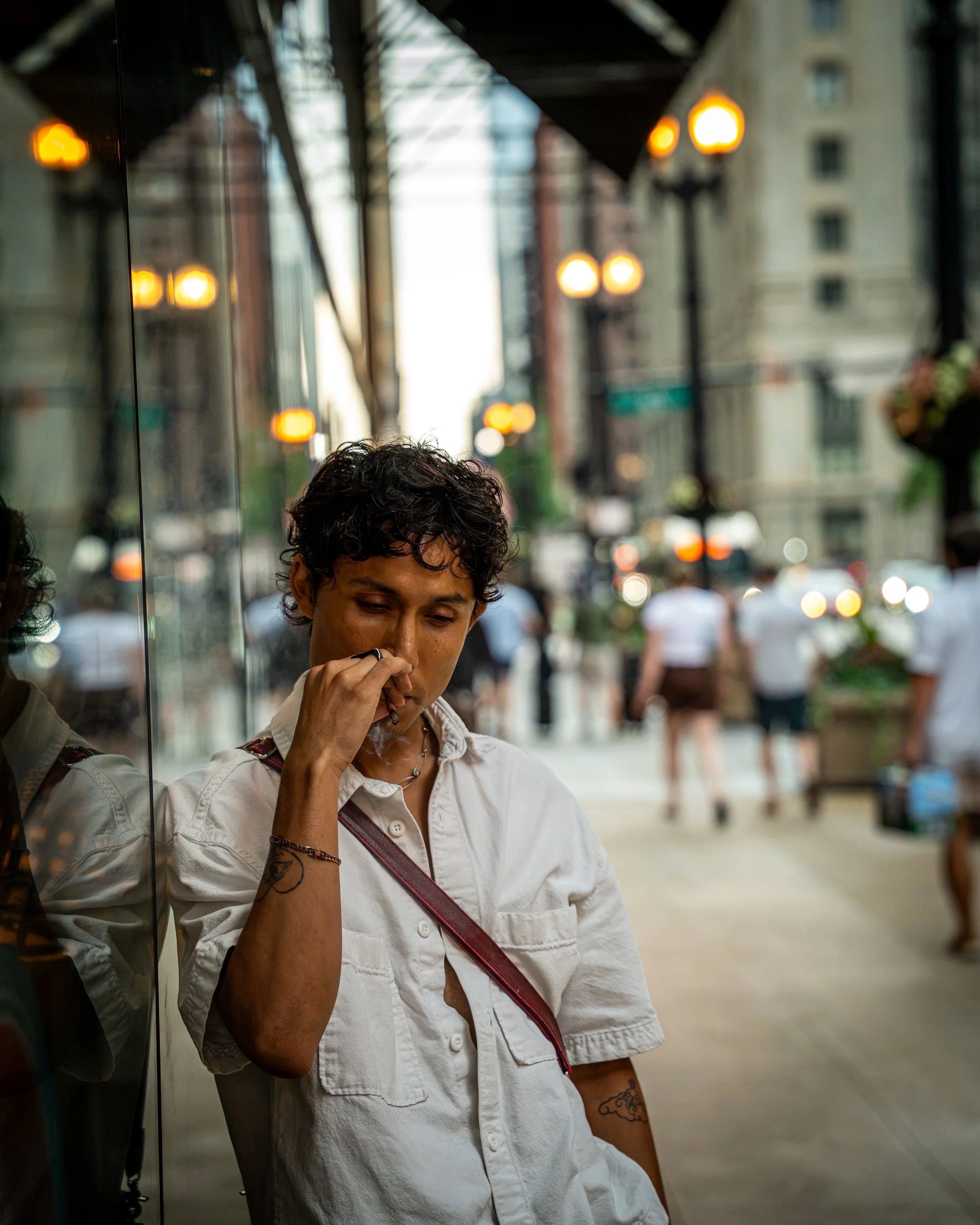 A person with short curly hair, wearing a white short-sleeved shirt, standing on a city street at dusk, talking on a phone. Reflection of the person can be seen in a window beside them. Blurred city buildings, streetlights, and pedestrians are visibl