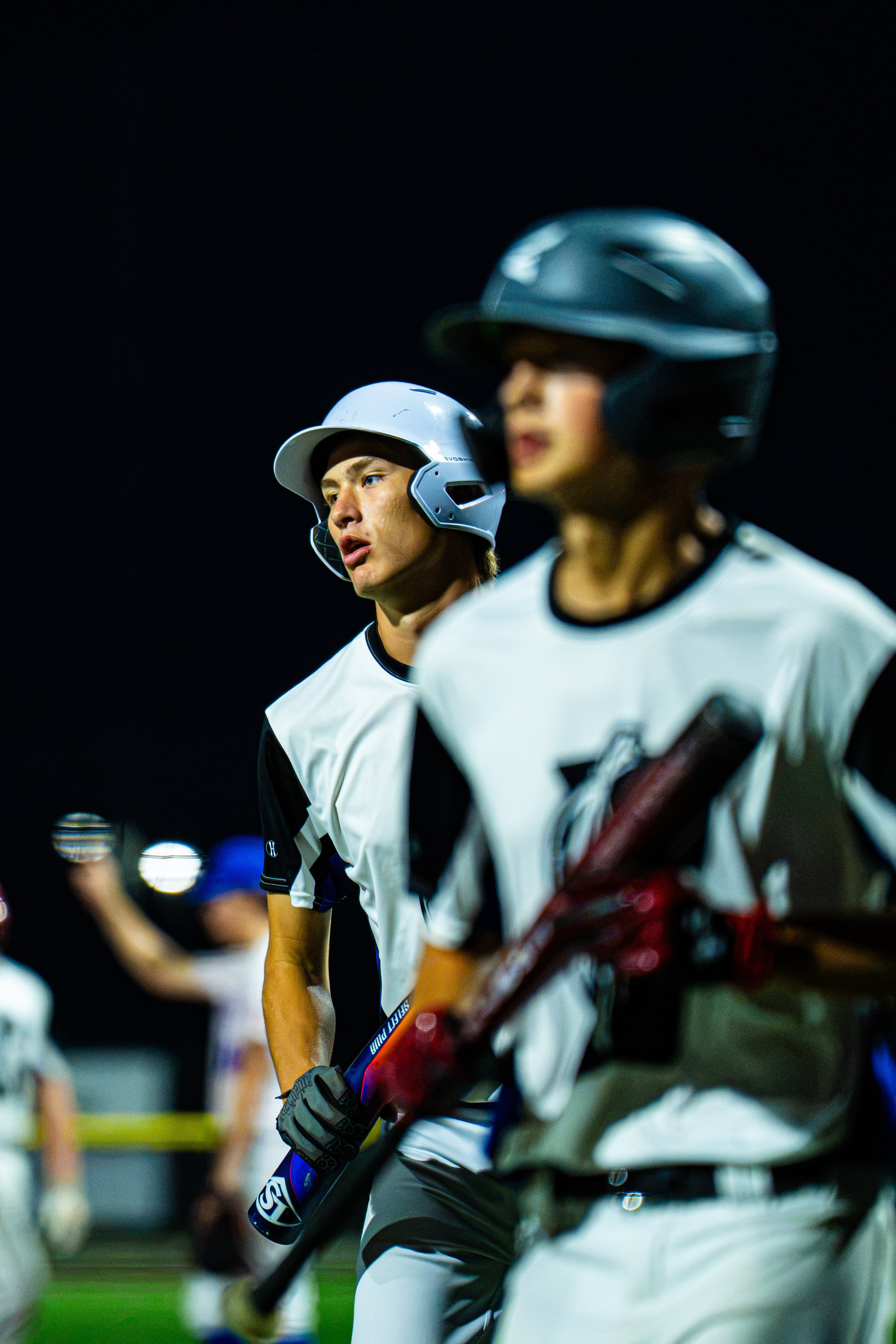Two baseball players in white jerseys and helmets, standing in a line during a night game or practice. The focus is on the player in the background with a serious expression, holding a bat, while the player in the foreground with a blurred face is al