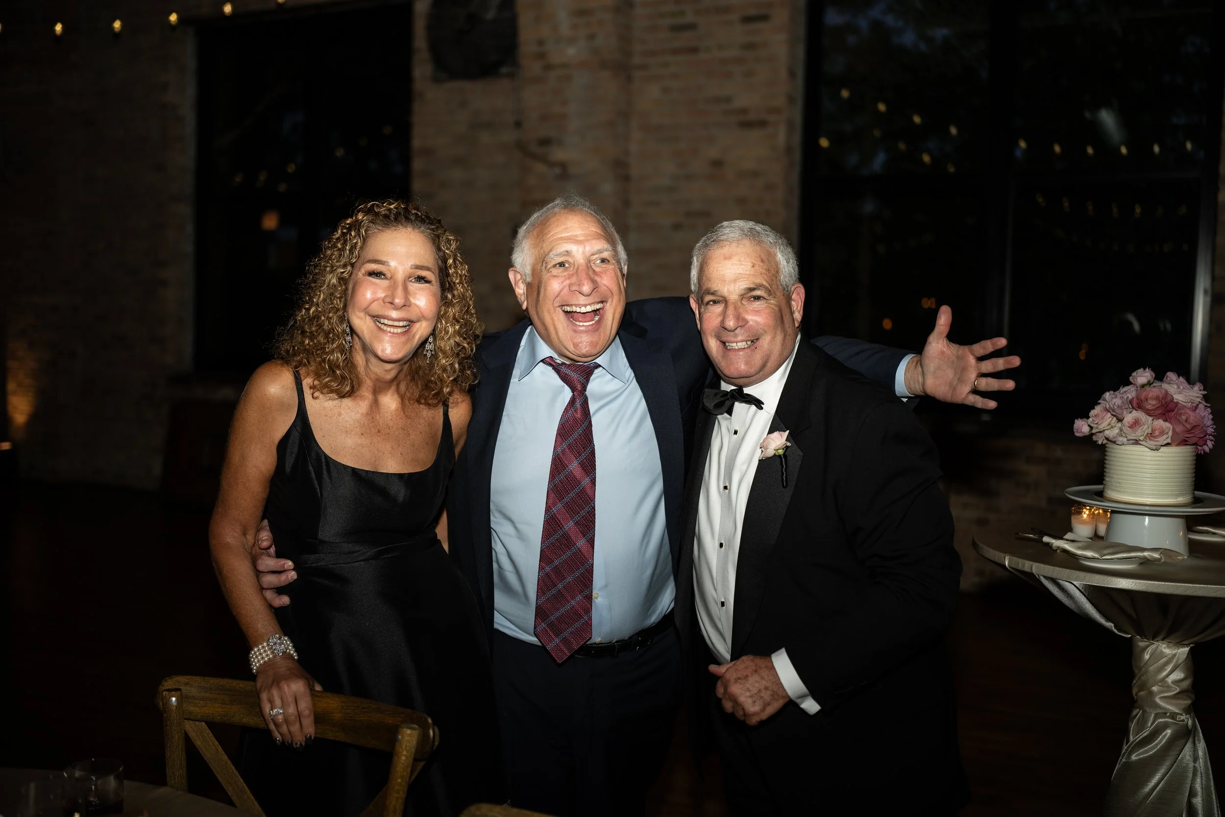 Three smiling people at a celebration, including a woman in a black dress and two men in formal attire, with a wedding cake and pink flowers on the table in front of them.