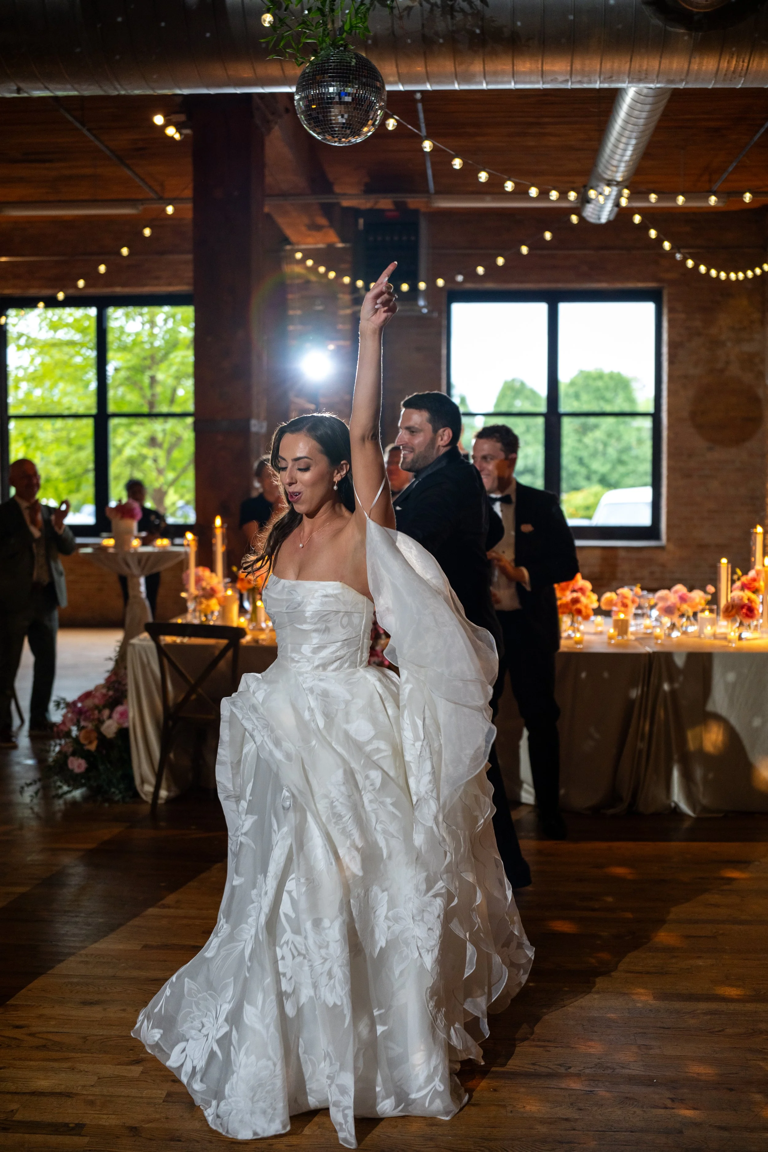 A bride in a white wedding dress is dancing with a man at an indoor wedding reception. The bride has her arm raised and is smiling. The background features large windows, string lights, and tables with floral centerpieces.