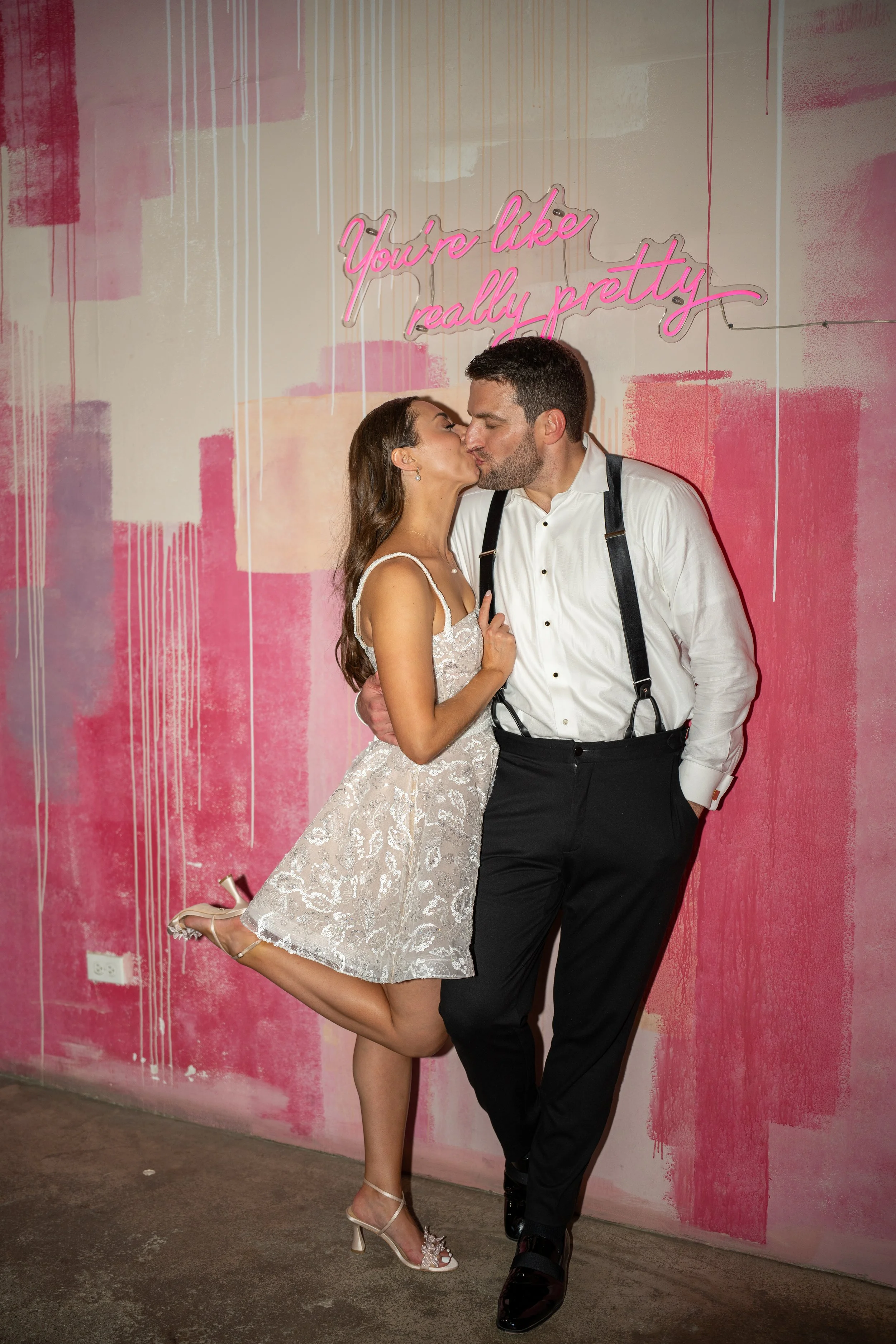 A couple kissing in front of a pink and white painted wall with a neon sign that reads 'You're like really pretty'.