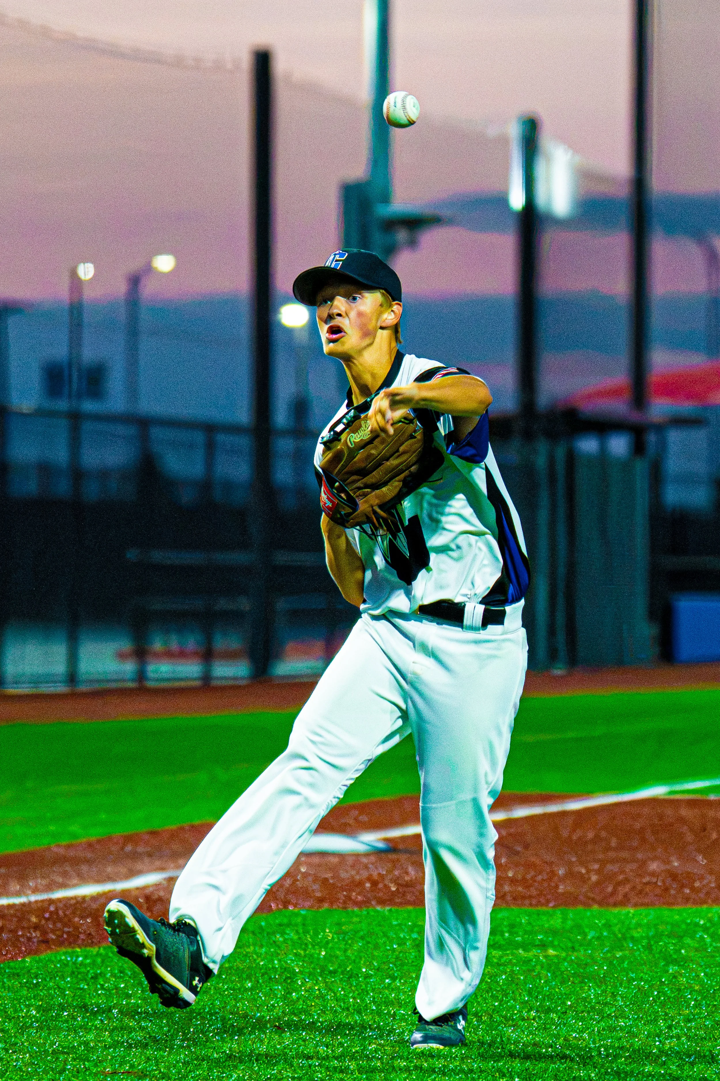 A baseball player is pitching on a baseball field during sunset. The player is in motion with his right leg raised and his arm extended, with a baseball in mid-air. The scene features a vibrant sky with pink and purple hues and is surrounded by field