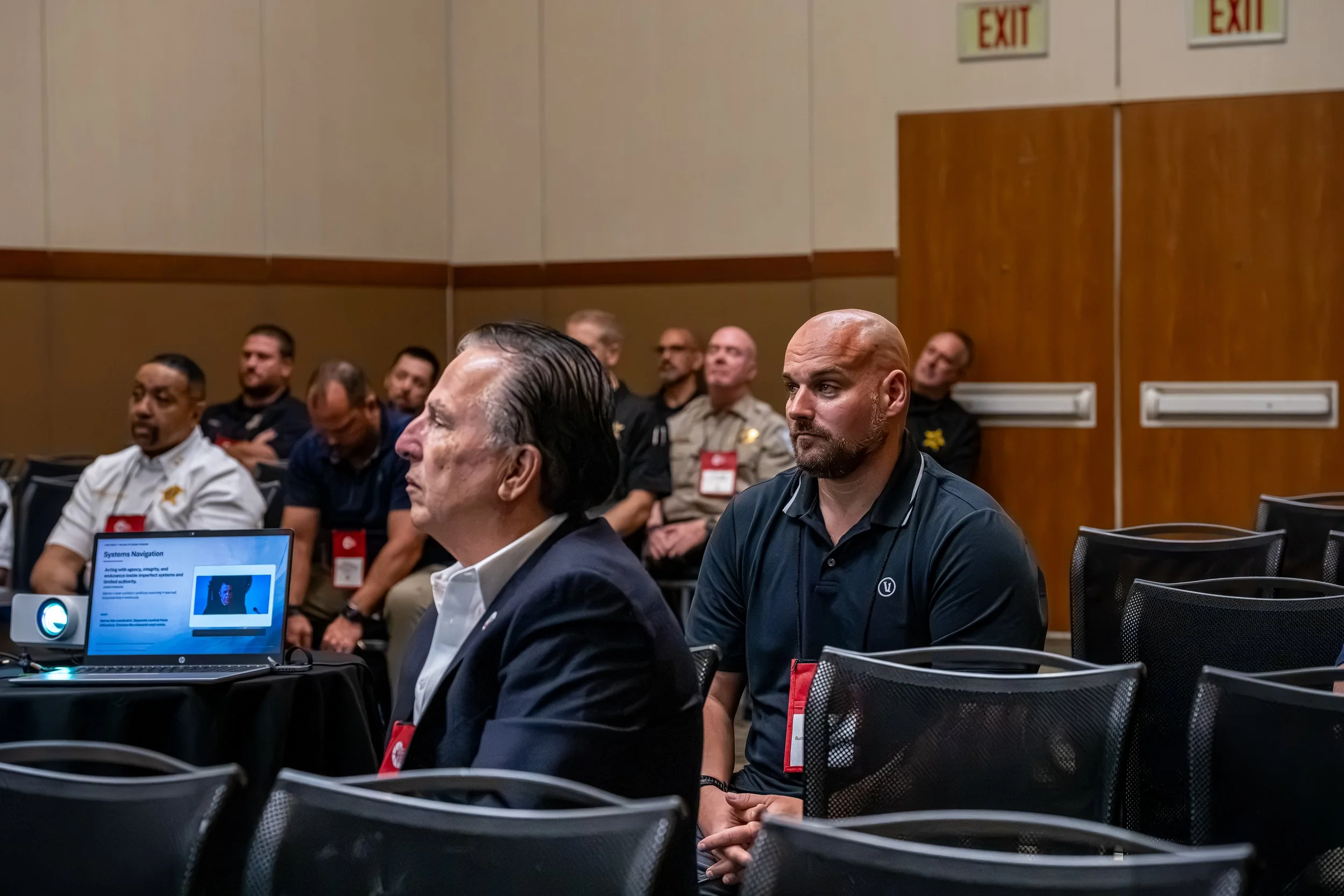 Attendees at a conference listening to a presentation, with a laptop showing a slide titled 'Systems Navigation', in a room with beige walls and exit signs.