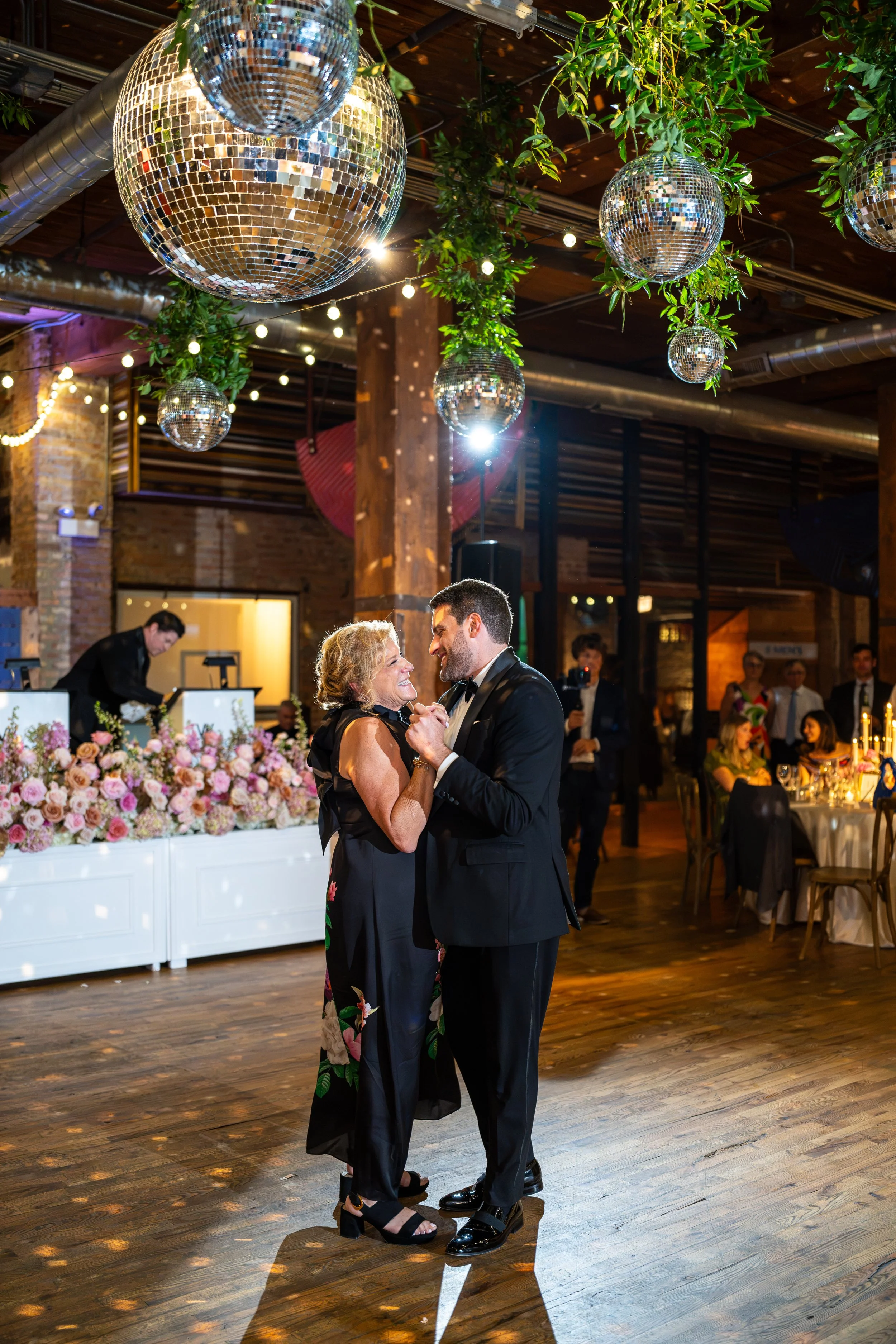 A couple dancing closely at a wedding reception under disco balls and string lights in a rustic, decorated venue with floral arrangements.