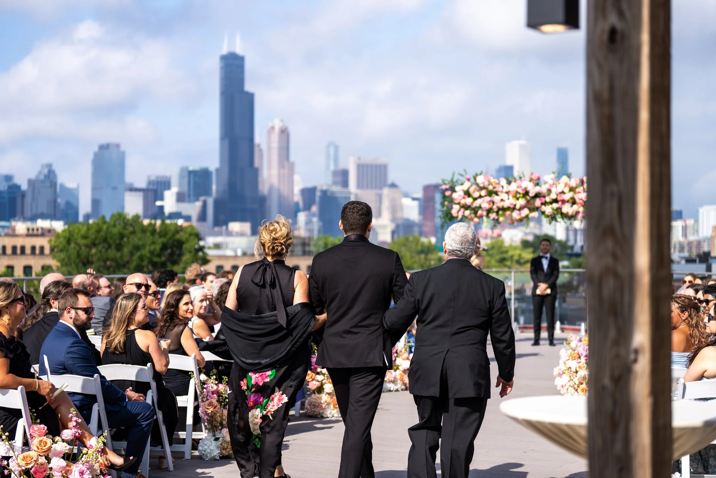 A wedding ceremony outdoors with guests seated and waiting, city skyline in the background, and some people walking down the aisle.