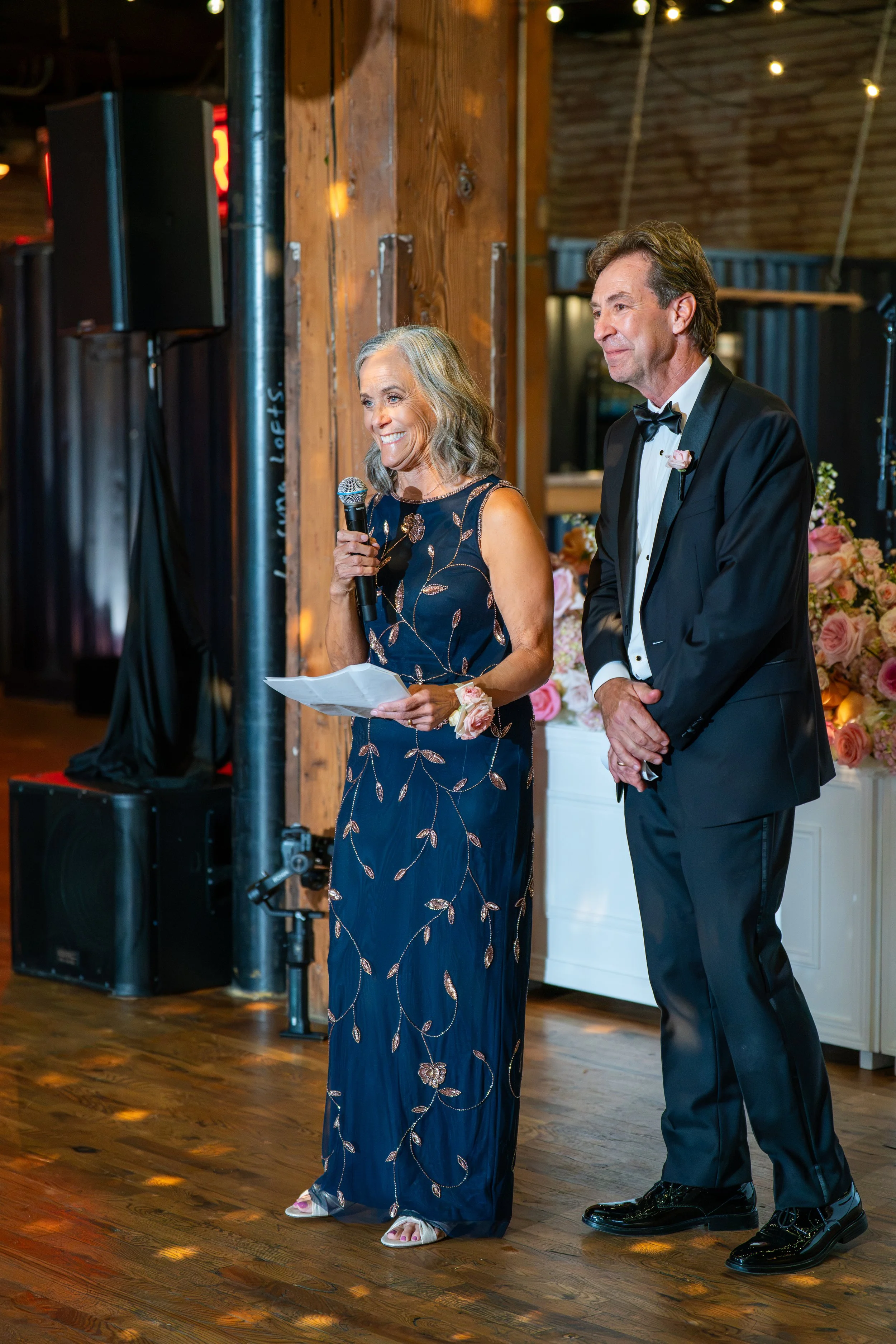 An older woman in a navy blue dress with gold embroidered leaves and flowers is smiling and reading from a paper, holding a microphone. Next to her, a man in a black tuxedo with a white shirt and black bow tie smiles and listens. They are at a weddin