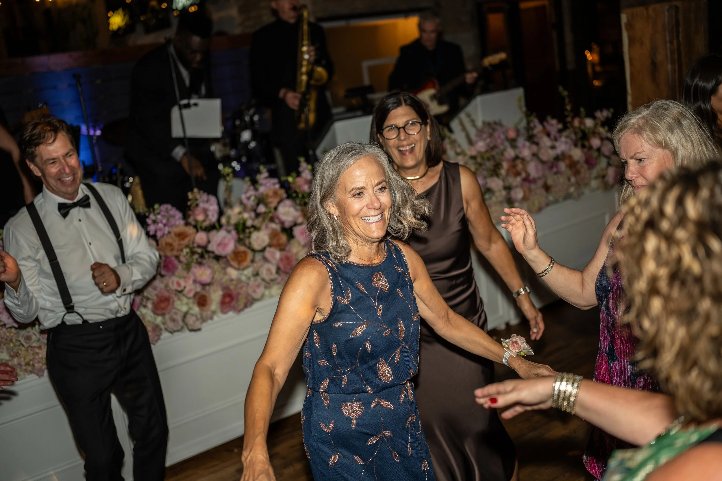 People dancing and celebrating at a wedding reception with floral decorations and live band playing in the background.