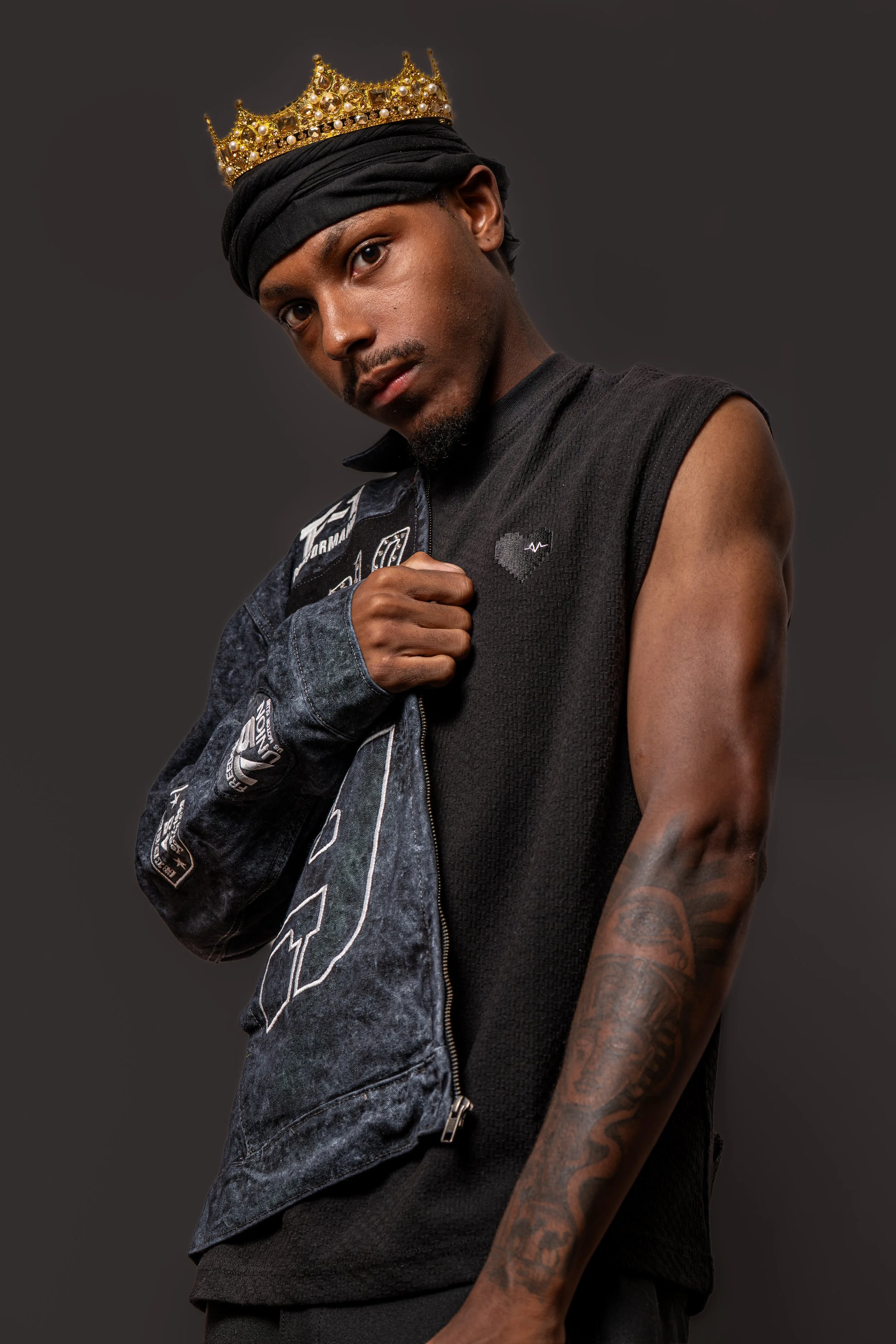 A young man wearing a black sleeveless shirt, a black headscarf, and a gold crown with pearls, posing with his right hand near his chest against a dark gray background. He has an arm tattoo visible.