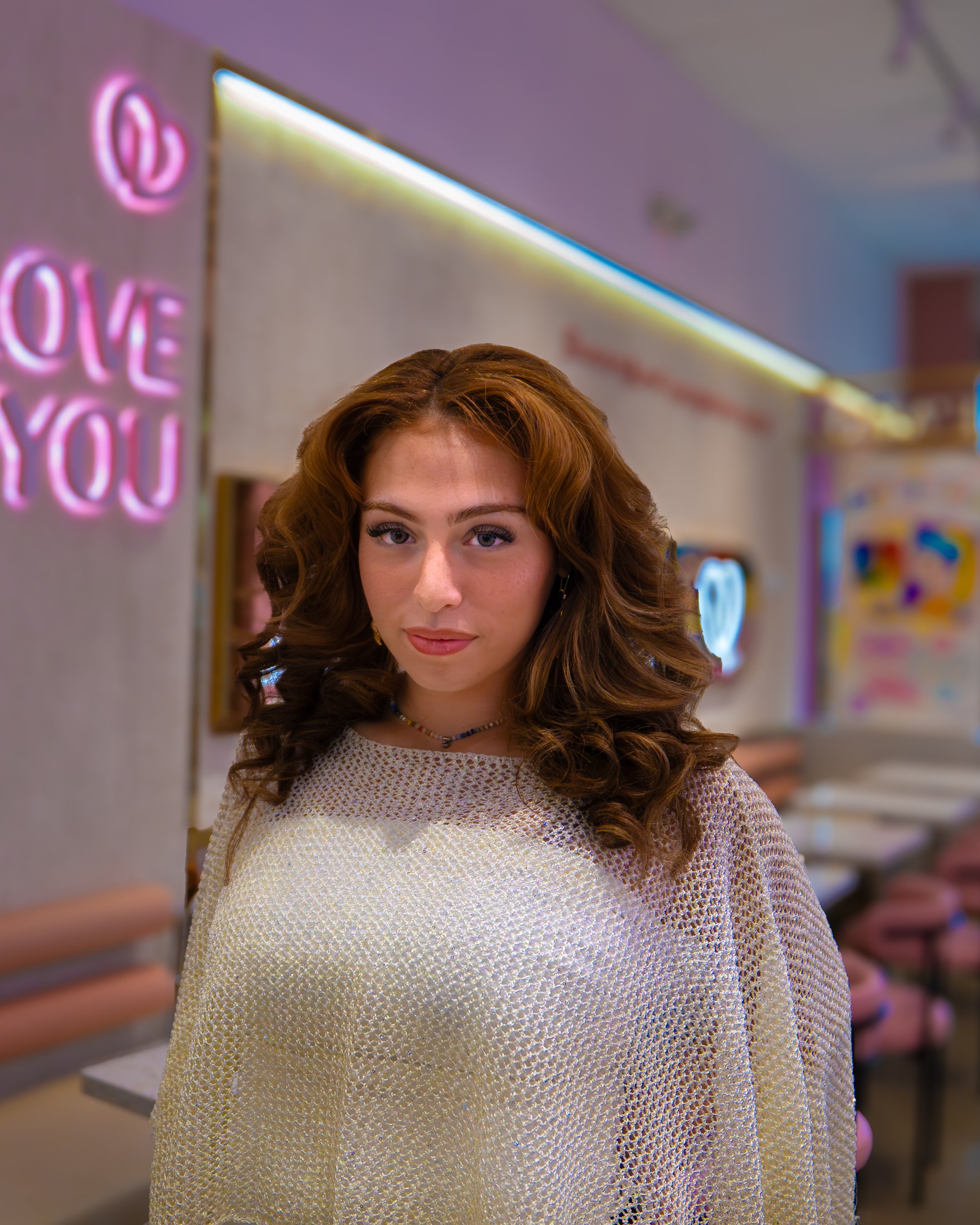 A young woman with red hair and blue eyes wearing a yellow crochet top and a beaded necklace, standing in a neon-lit space with pink and purple signs, including one that says 'LOVE YOU'.