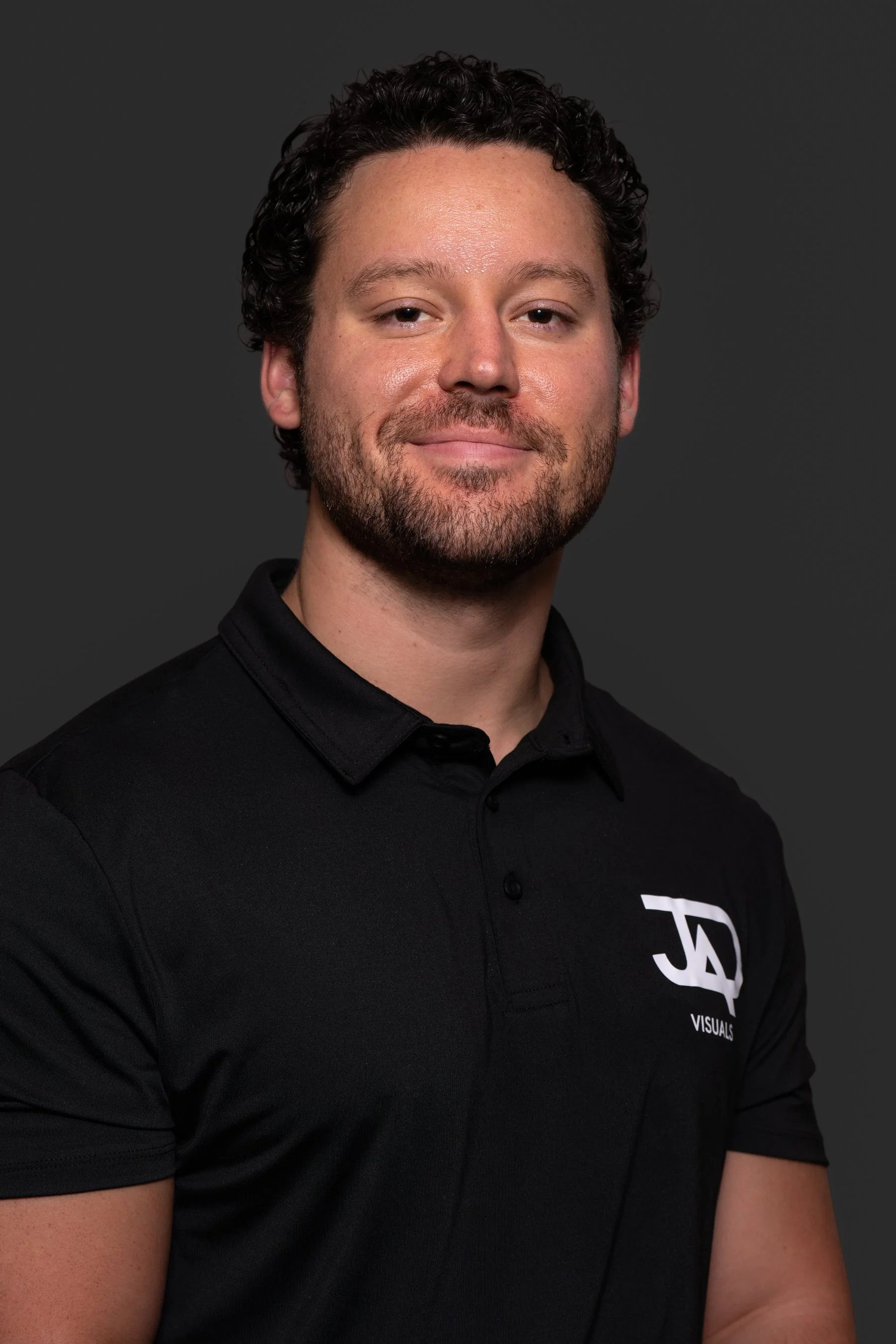 A man with curly dark hair and a beard wearing a black polo shirt with a logo on the chest, smiling softly against a dark background.