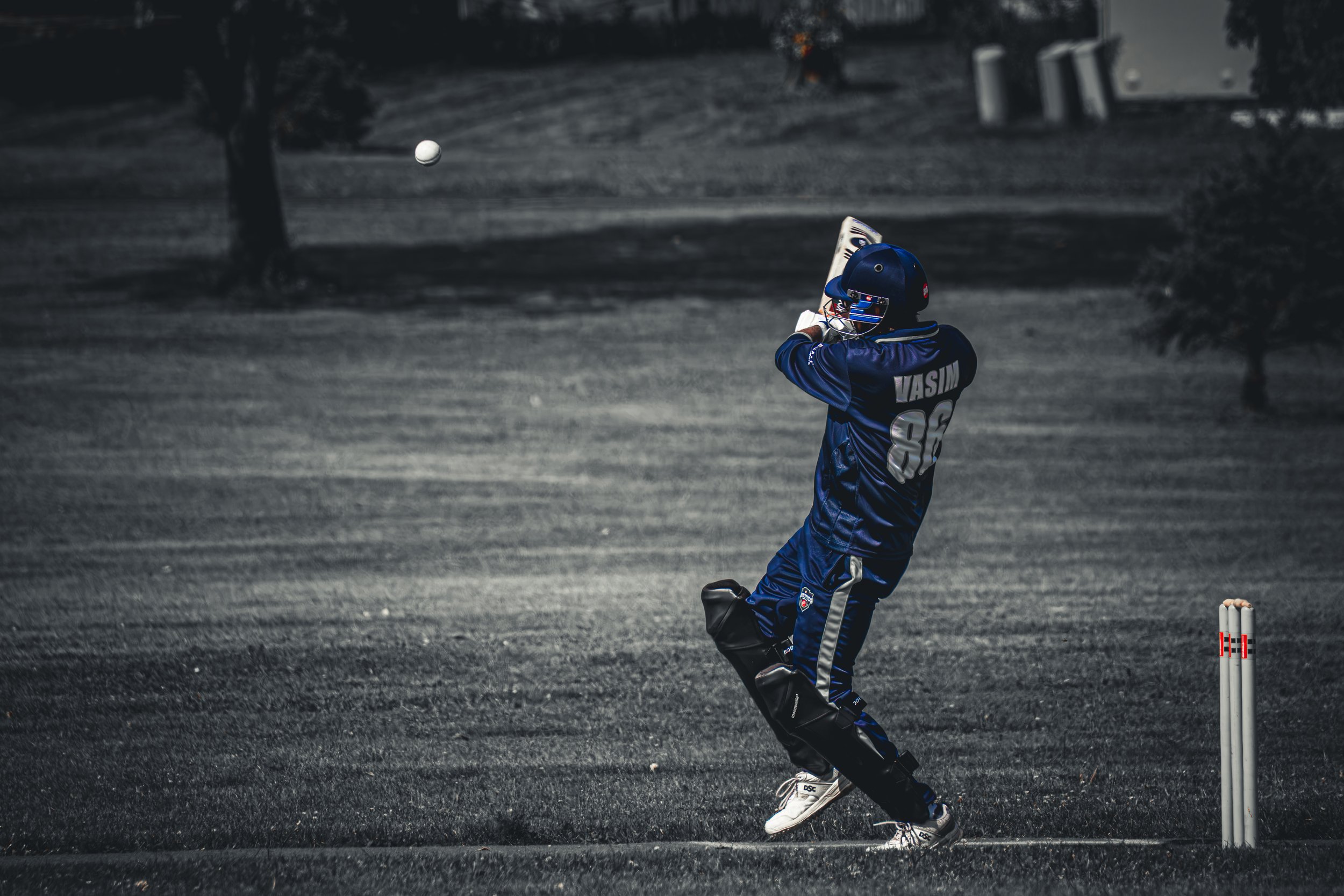 A young cricket player in blue uniform and helmet, playing cricket outdoors with a white ball mid-air, black sunglasses, and gloves, on a dark ground with trees and a building in the background.