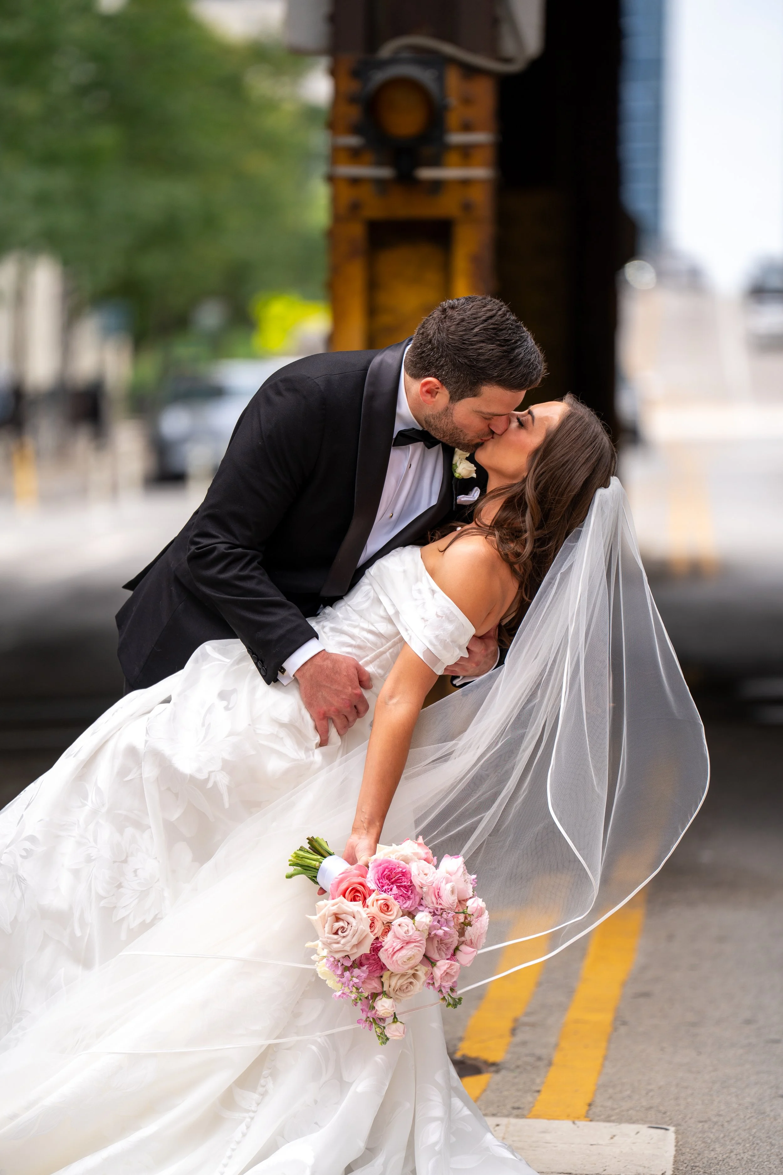 A newlywed couple sharing a kiss outdoors. The groom, in a black tuxedo with a bow tie, is holding the bride, who is wearing a white wedding gown and veil. She is holding a pink and white bouquet of flowers.