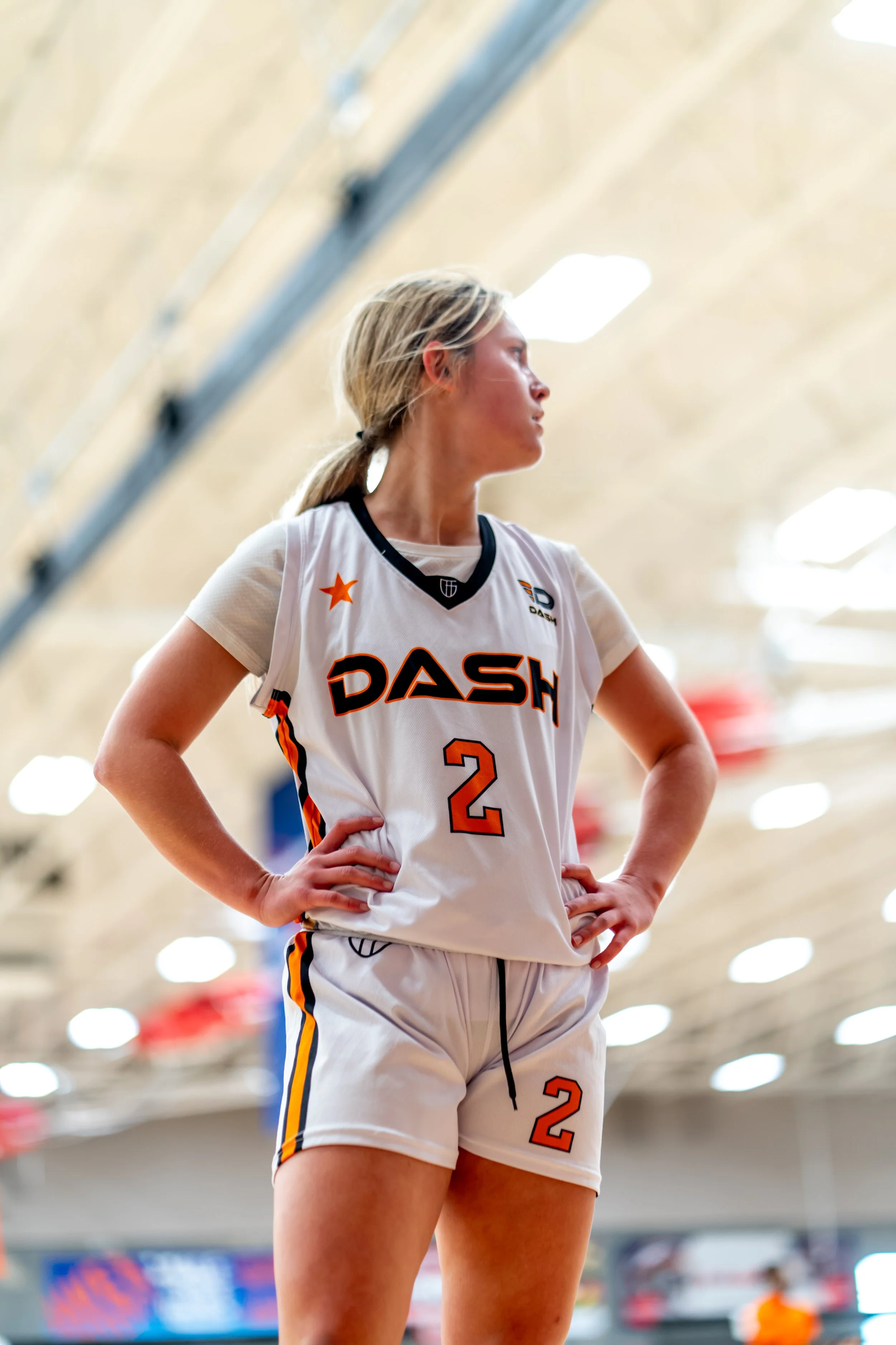 A female basketball player wearing a white uniform with orange and black accents stands on the court with her hands on her hips. She has blonde hair tied back and is looking to the side.