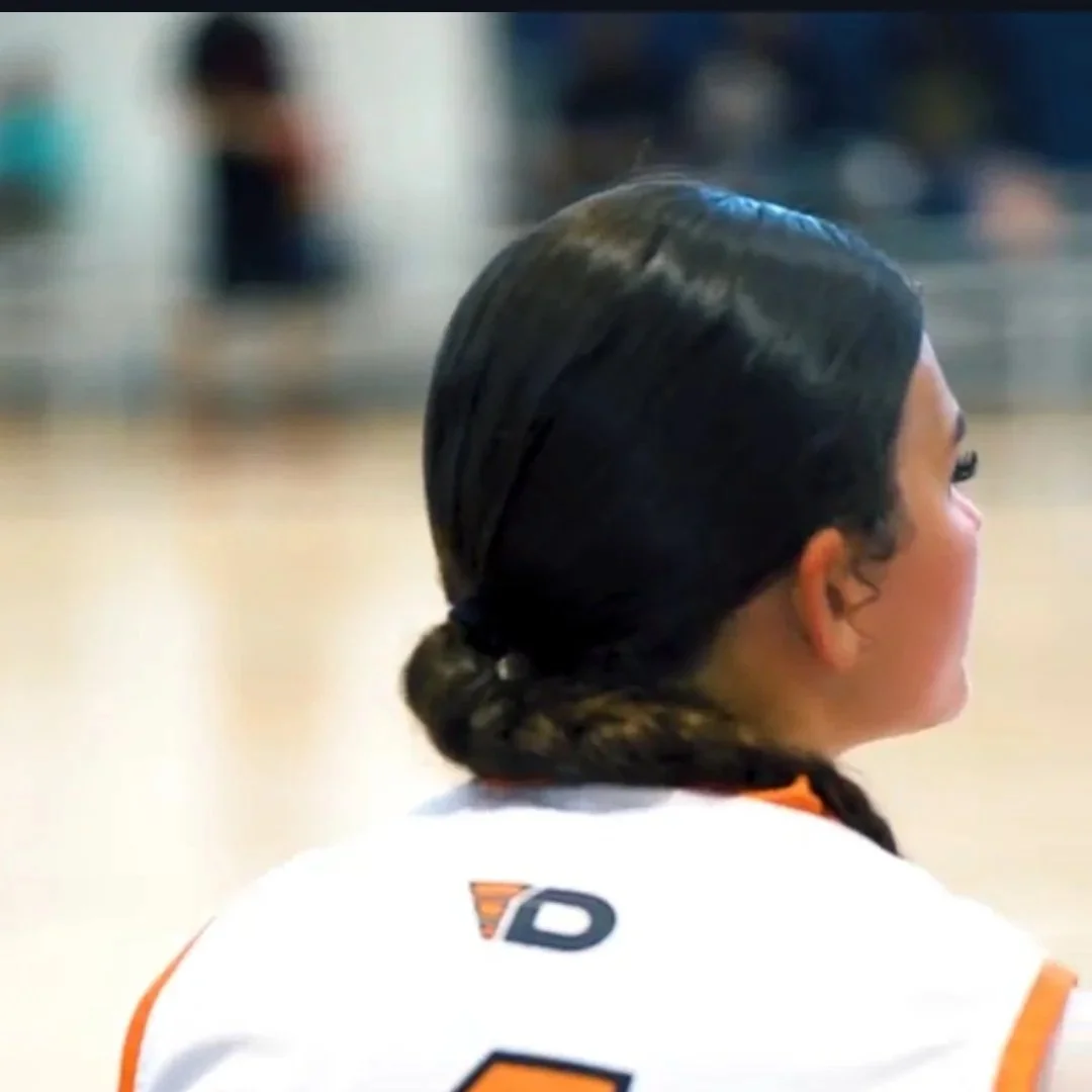 A woman with dark hair in braided bun, wearing a white sports jersey with orange accents, sitting on a sports court.