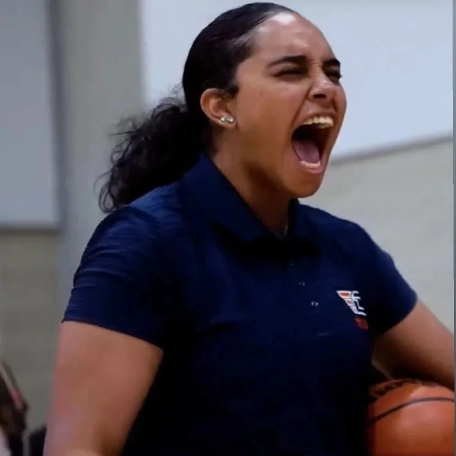 Woman with dark hair, yelling passionately, holding a basketball, wearing a navy blue shirt with a logo.