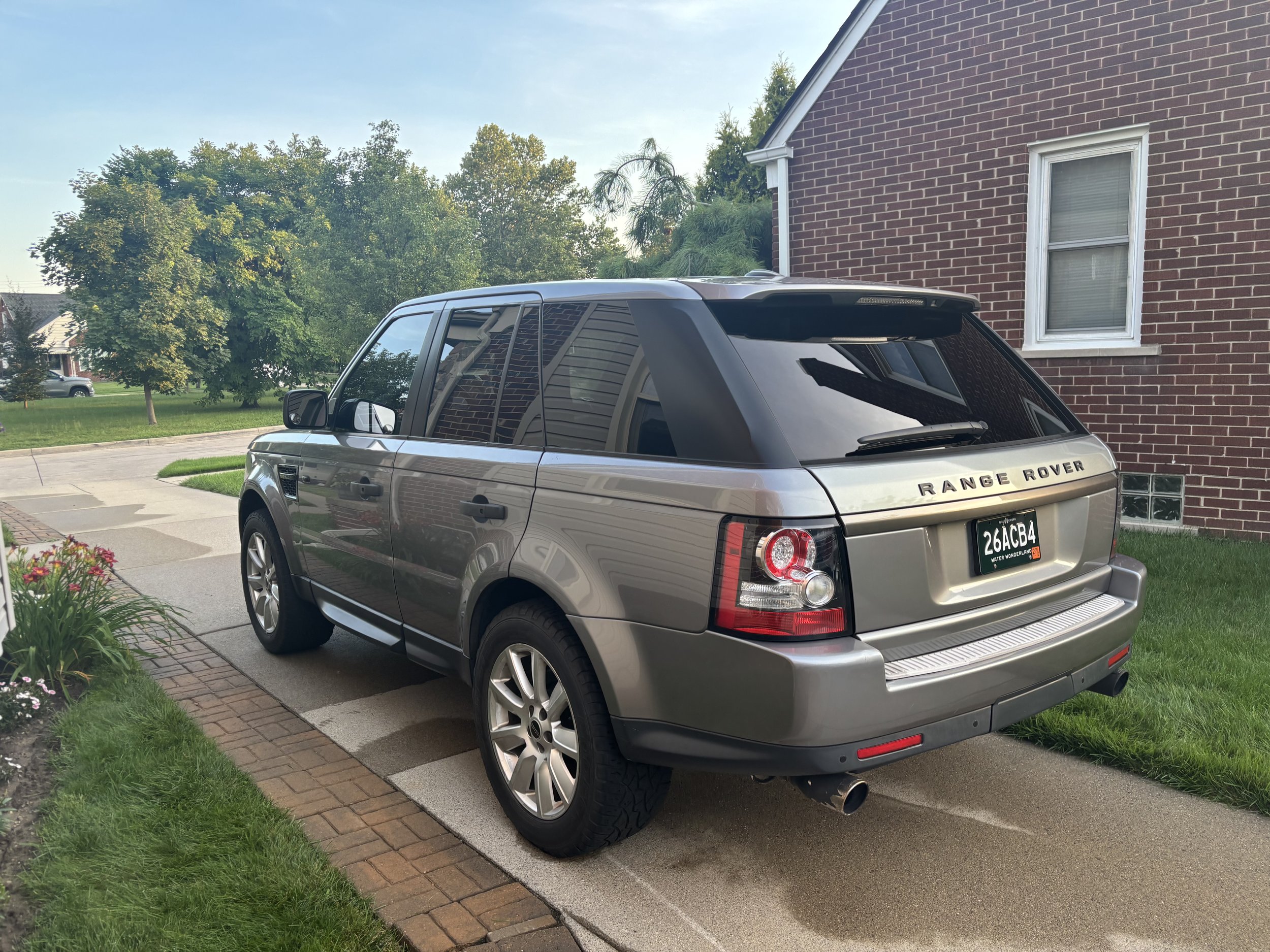 A silver Range Rover parked in a residential driveway, with a brick house and green trees in the background.