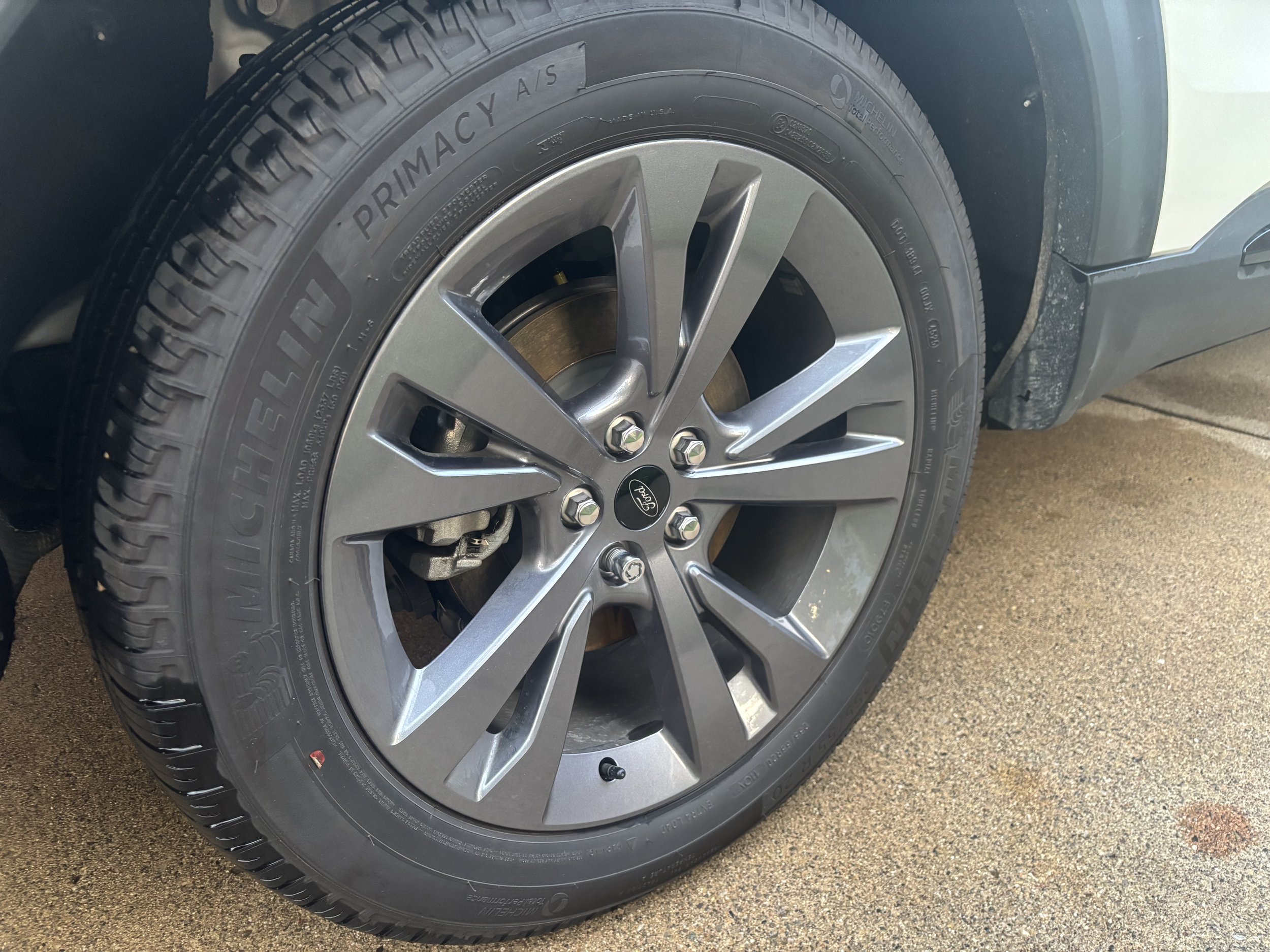 Close-up view of a car tire with a silver alloy wheel. The tire has the brand name 'PRIMACY A/S' visible. Part of the car's body and a concrete surface are also visible.