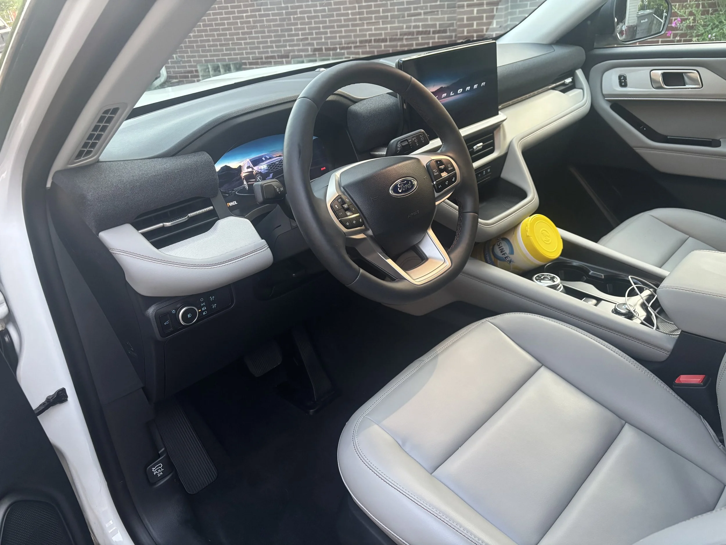 The interior of a modern Ford vehicle showing a steering wheel, dashboard with digital display, a large central screen, and light-colored leather seats. A can of disinfecting wipes and a container are in the cup holder area.
