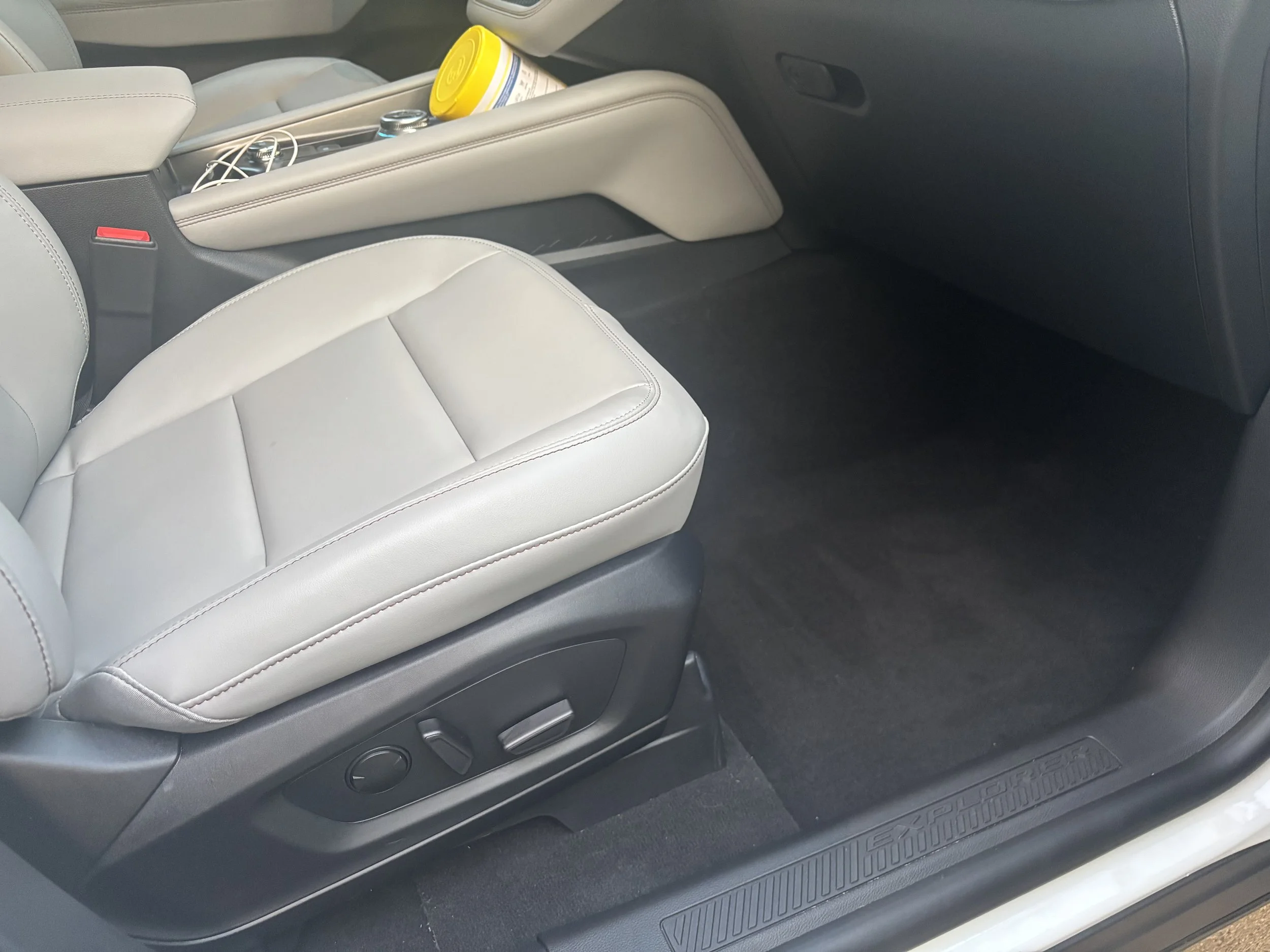 Inside a vehicle showing the back seat, with a gray leather headrest, a center armrest with a container and some wires, and a black carpeted floor with a textured floor mat.