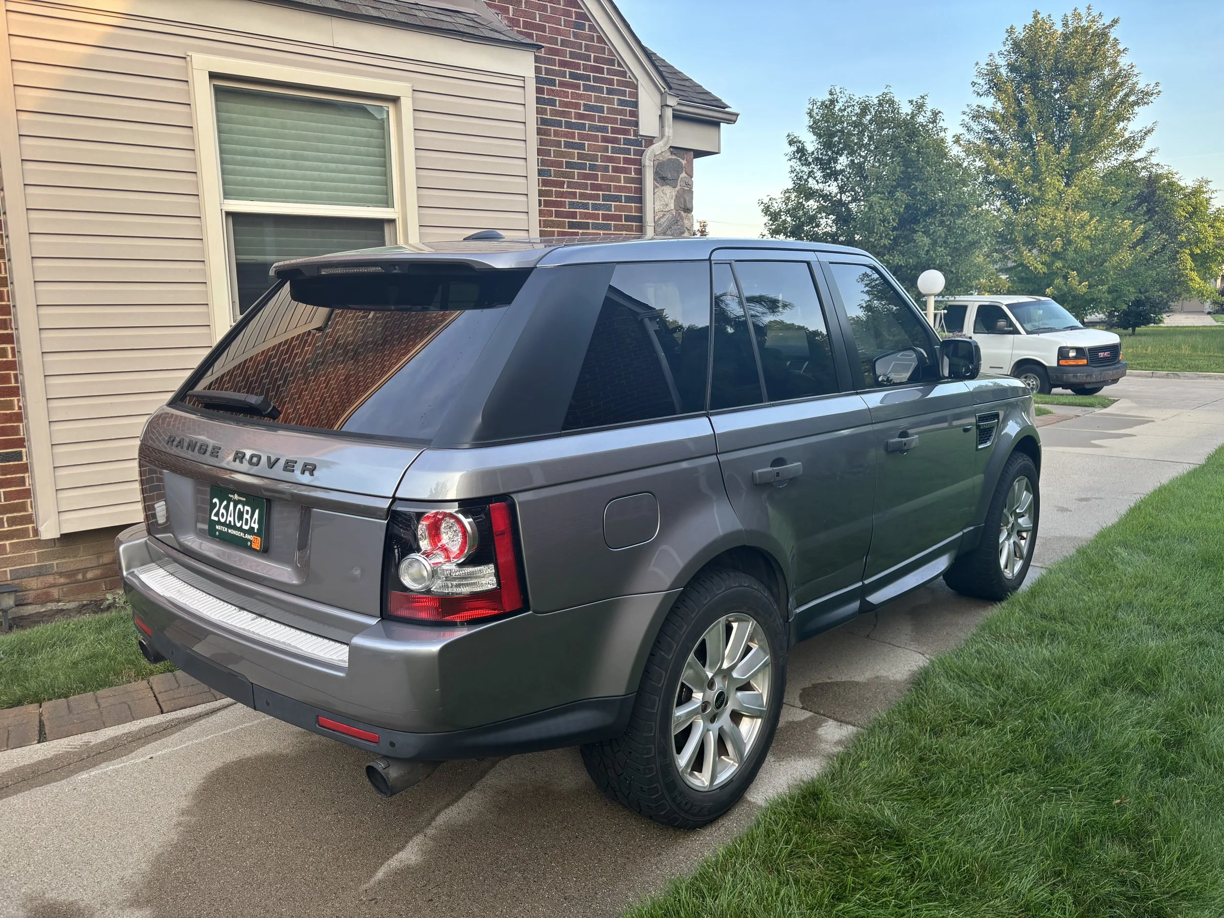 A silver Range Rover parked on a driveway next to a house with beige siding and brick wall. In the background, there is a gray GMC van, green trees, and a clear blue sky.