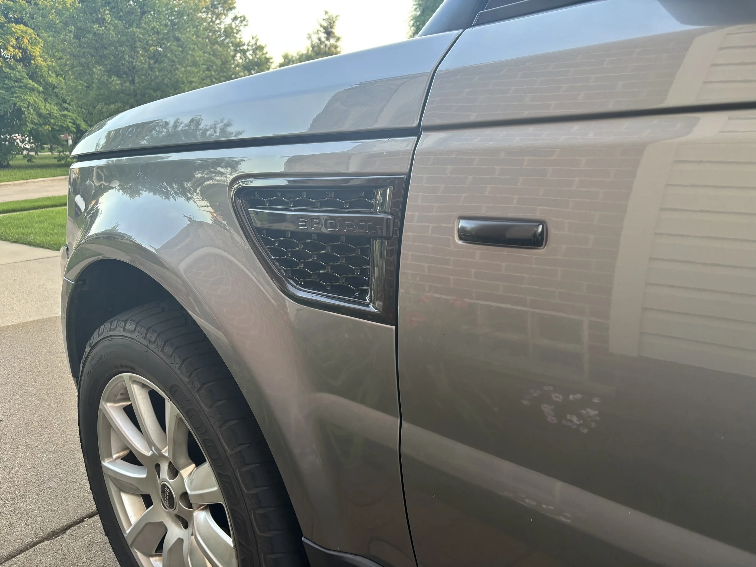Close-up of the front right side of a silver or gray vehicle, showing the headlight, part of the grille with the word 'SPORT', a black vent, and the tire with alloy wheel, with a background of green trees and a sidewalk.