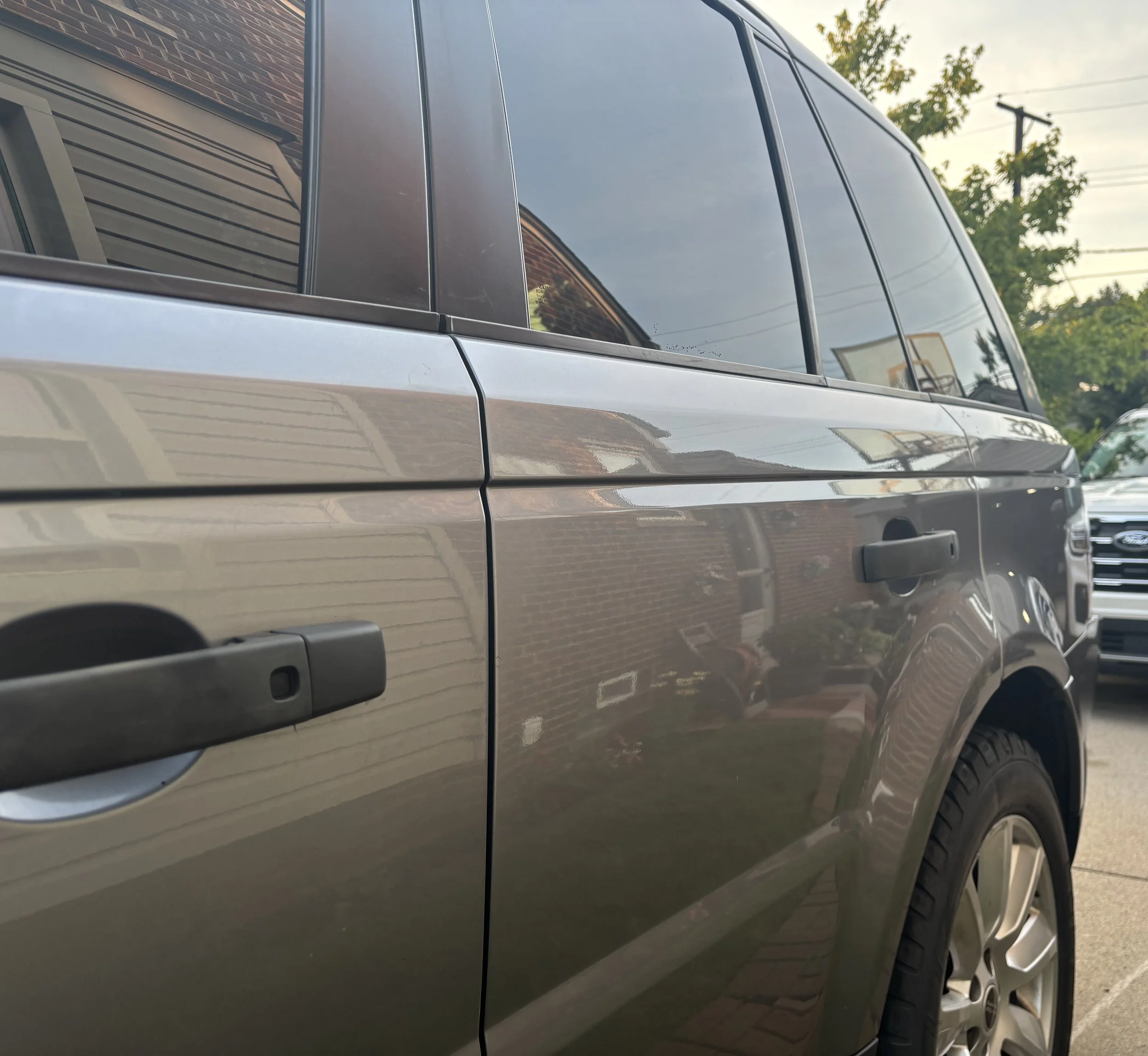 Close-up of a grey SUV parked outdoors, showing part of the driver's side door, window, and rearview mirror with trees and power lines in the background.