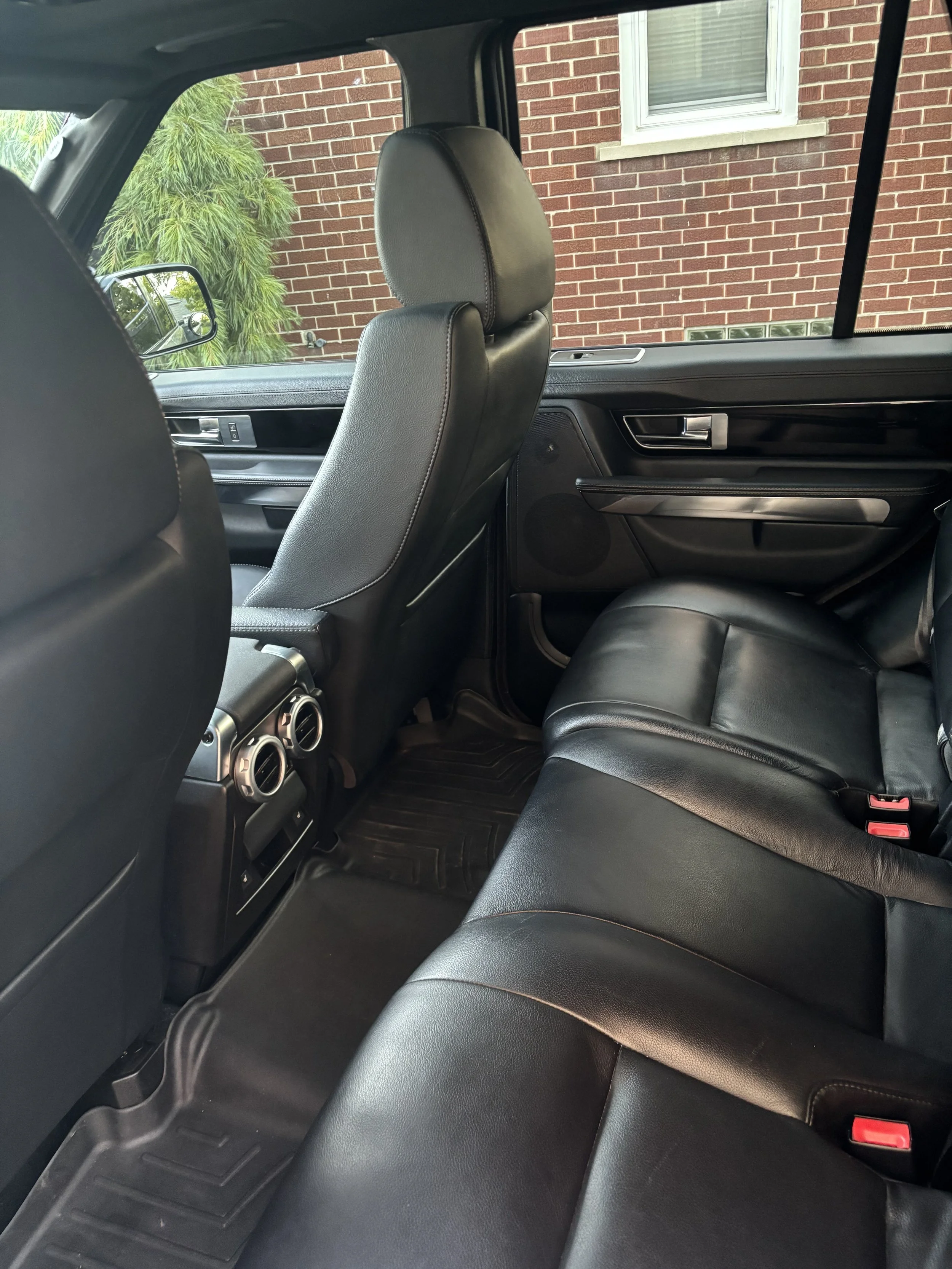 The interior of a black SUV with leather seats, showing the back seat, front passenger seat, and part of the door, with a brick house and window visible through the window.