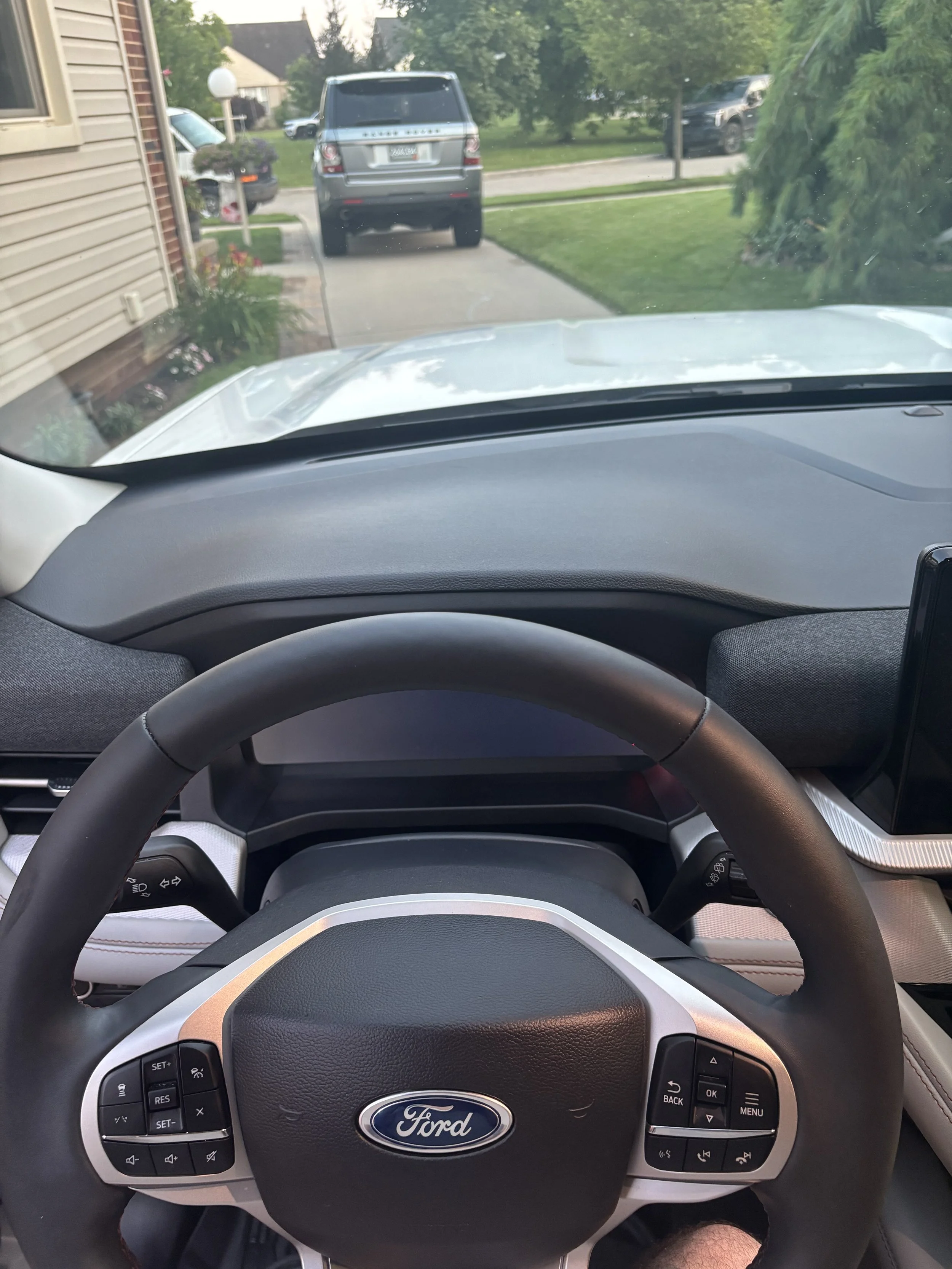Inside view of a Ford vehicle dashboard, with steering wheel and control buttons, looking out through the windshield at a driveway with parked cars and a grassy yard with trees.