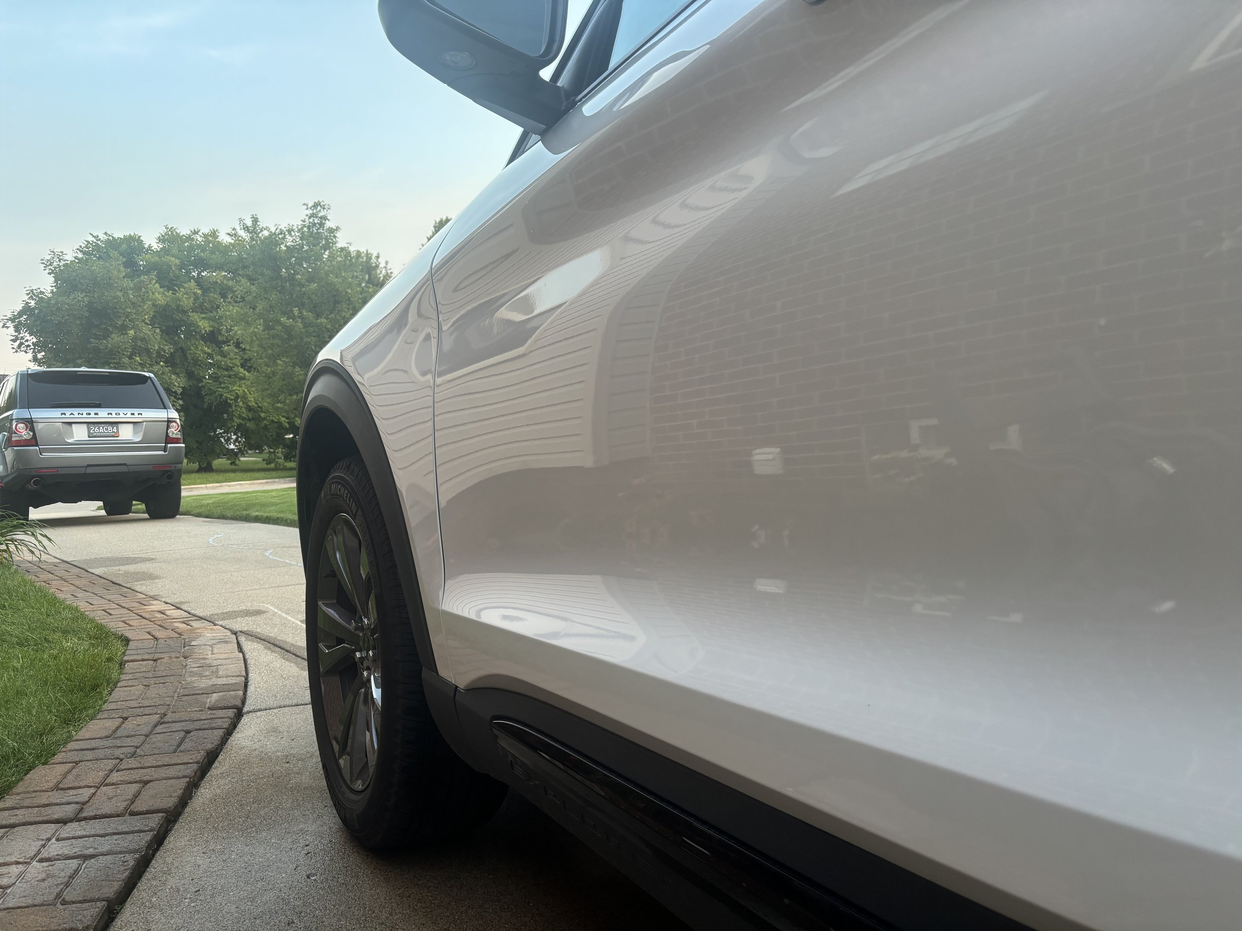 Close-up of a silver car parked on a driveway, with another car visible in the background and trees beside the driveway.