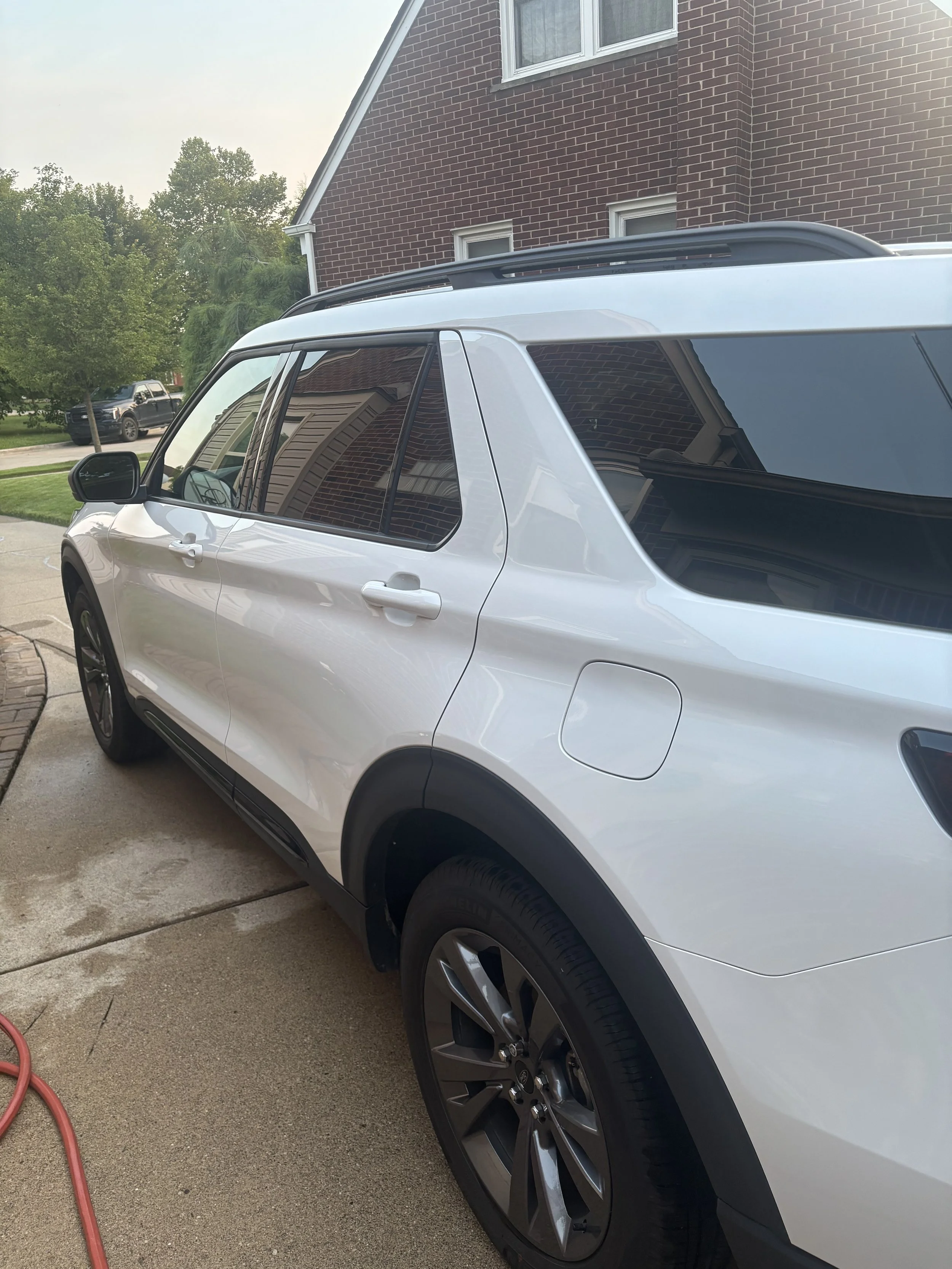 A white SUV parked outside a house with a brick exterior. The house has several windows and a gable roof. A red garden hose lies on the driveway near the vehicle. Trees and another parked vehicle are visible in the background.