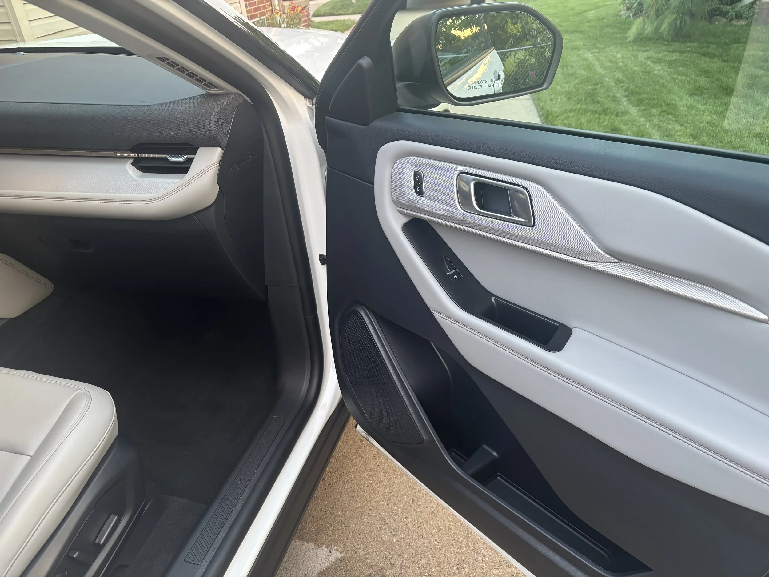 Photo of the inside of a vehicle showing the front passenger side door with black and white interior design, window controls, and a side mirror reflecting a lawn and houses outside.