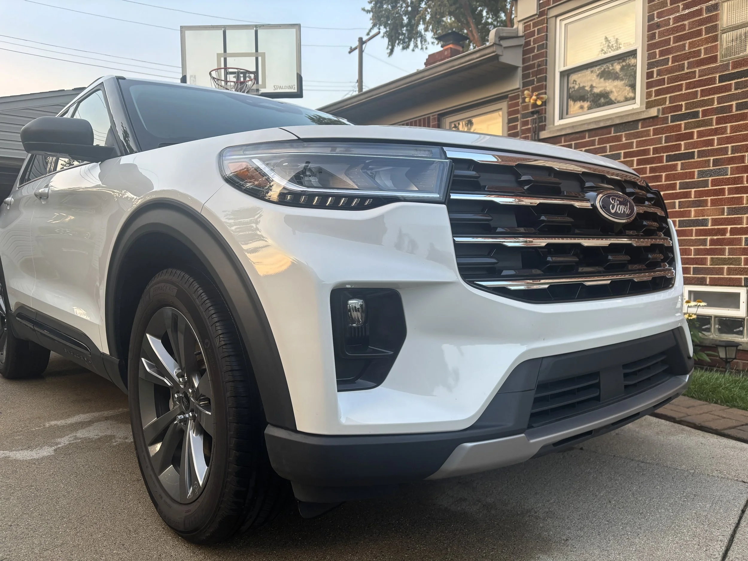 A white Ford SUV parked on concrete driveway in front of a brick house with a basketball hoop visible above the vehicle. There is a window with a butterfly decoration on the house and some plants visible nearby.