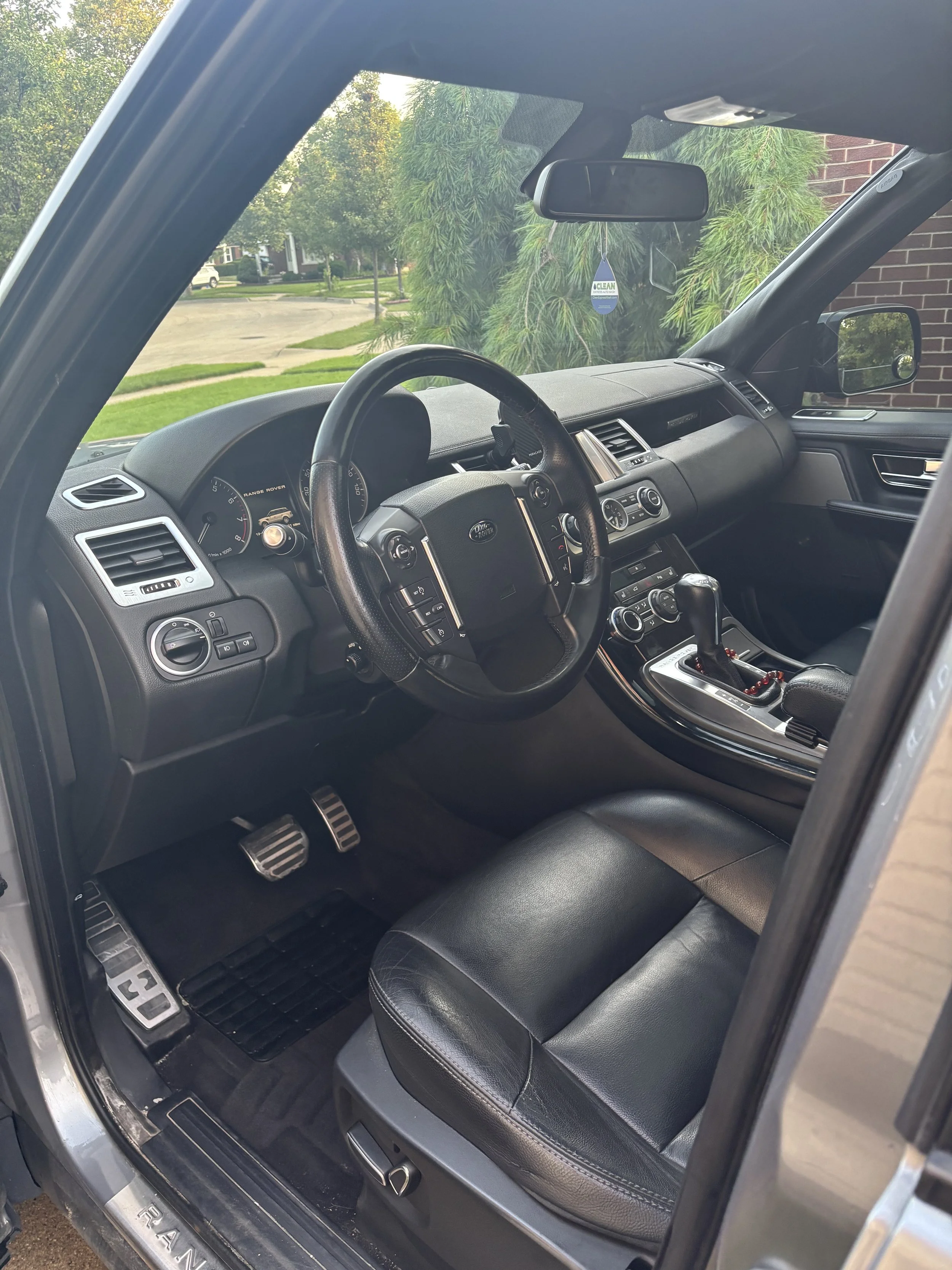 Interior of a Range Rover vehicle showing the dashboard, steering wheel, gear shift, and black leather seats, with a view of the outside greenery through the windows.