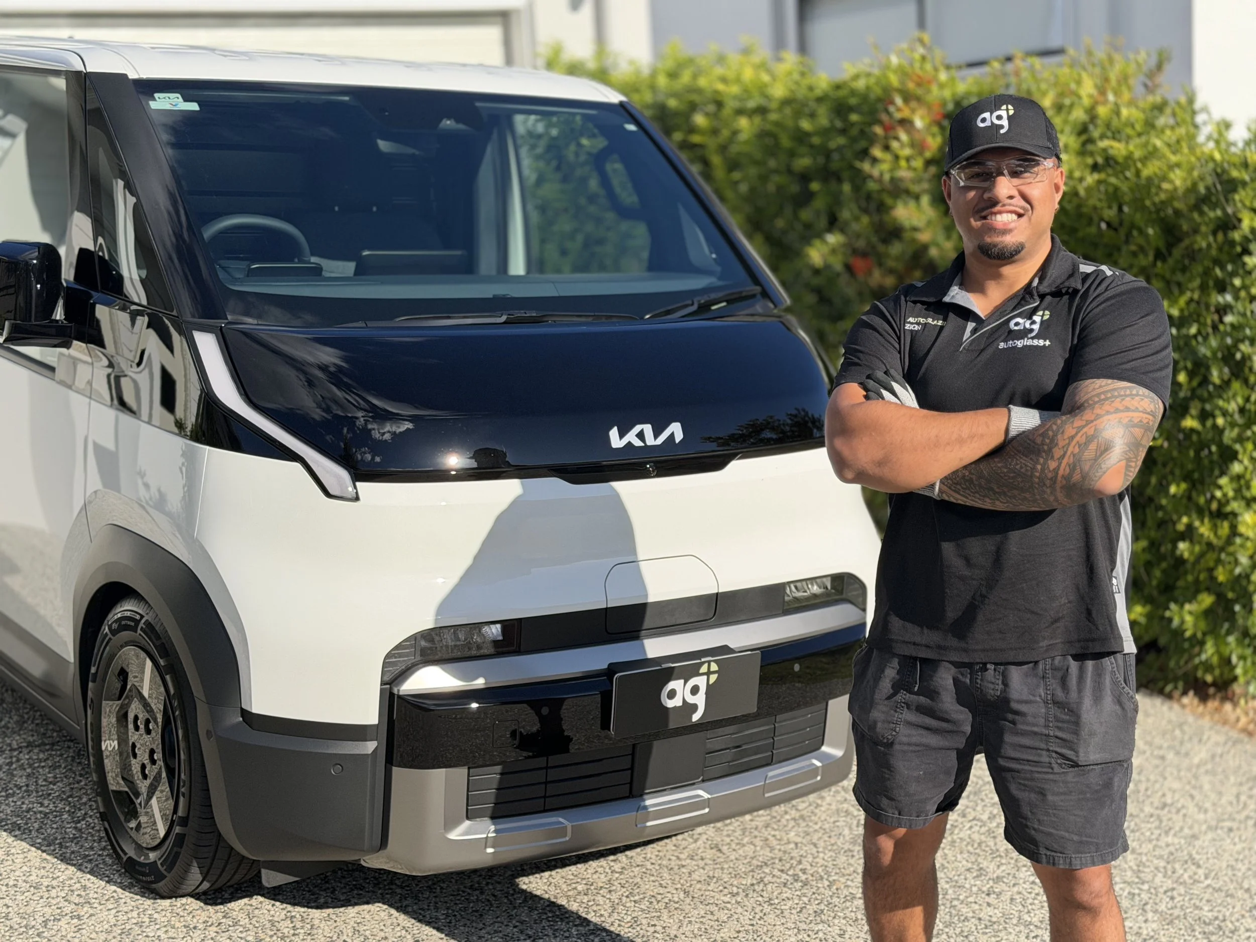 A man standing with arms crossed next to a white Kia electric vehicle outdoors on a sunny day with green bushes in the background.