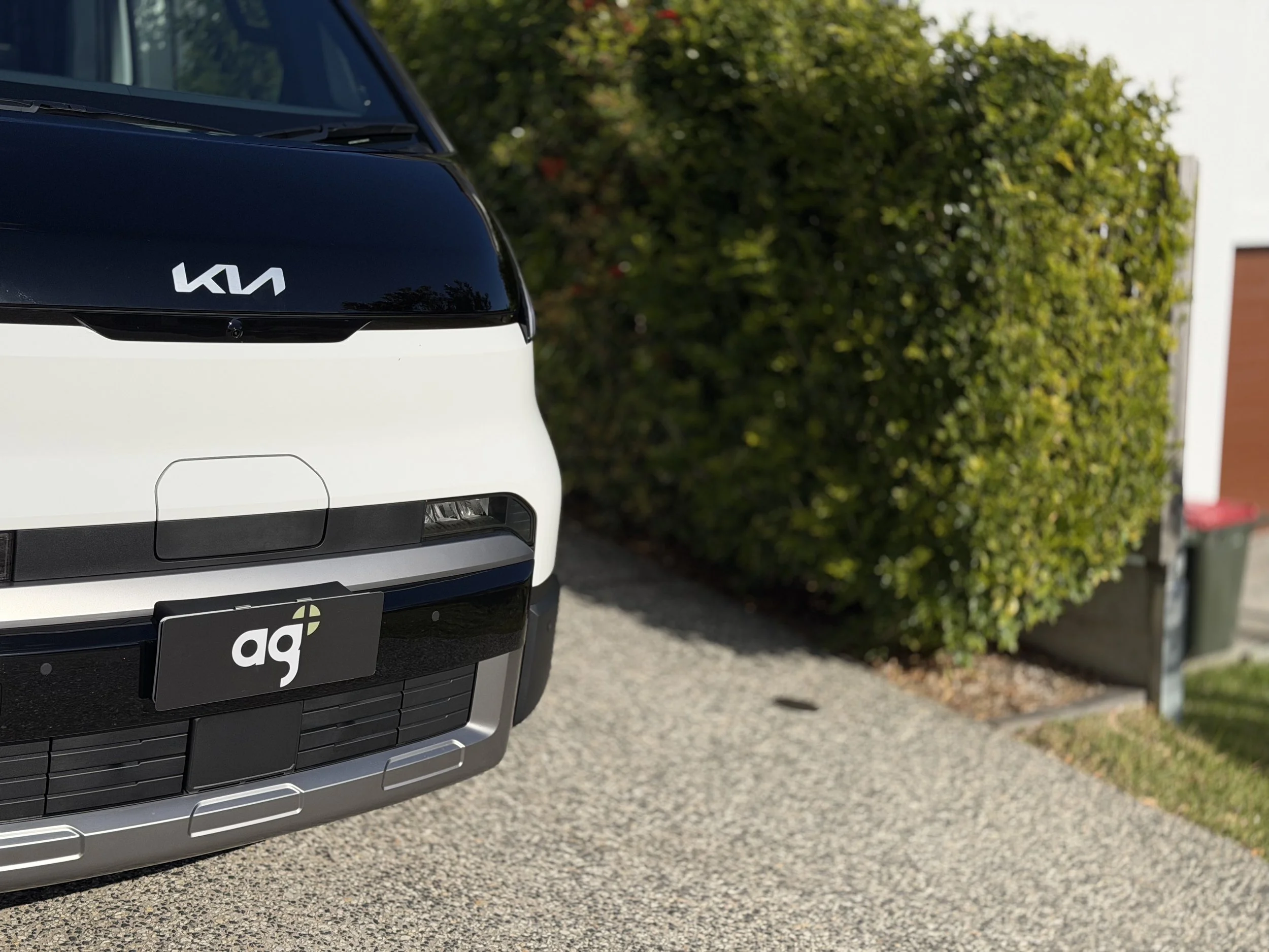 Close-up view of the front part of a white Kia vehicle parked outdoors in daytime, with a background of green bushes and a building.