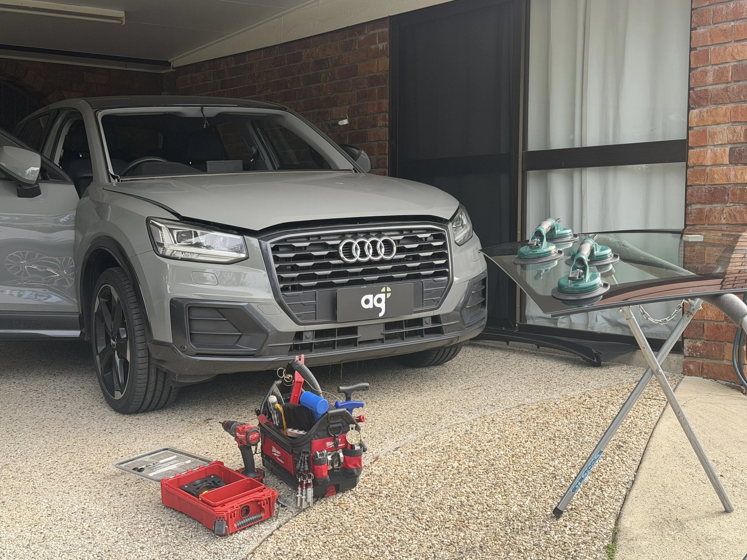A silver Audi SUV parked under a carport against a brick wall. In front of the car, there are toolboxes and tools, including a red Milwaukee toolbox, a cordless drill, and other hand tools on the ground. To the right, there is a glass-top table with green steam irons on it, and in the background, there is a window with white curtains.