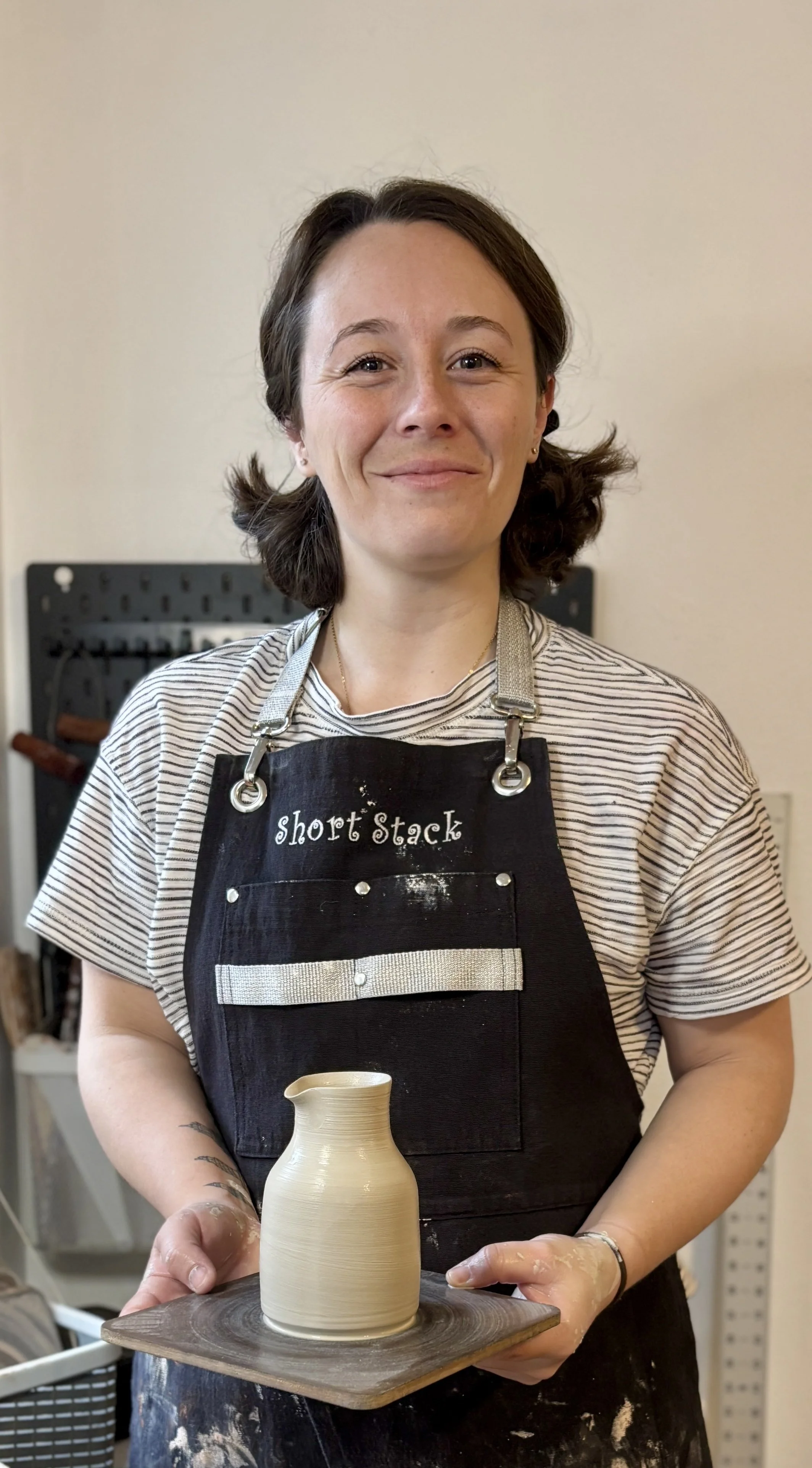 A woman smiles while holding a pottery piece on a wheel, wearing an apron that says "Short Stack".