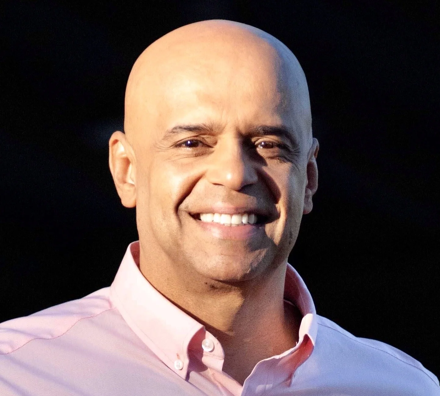Serge Fiankan (SF) headshot smiling with a pink button down shirt on a black background