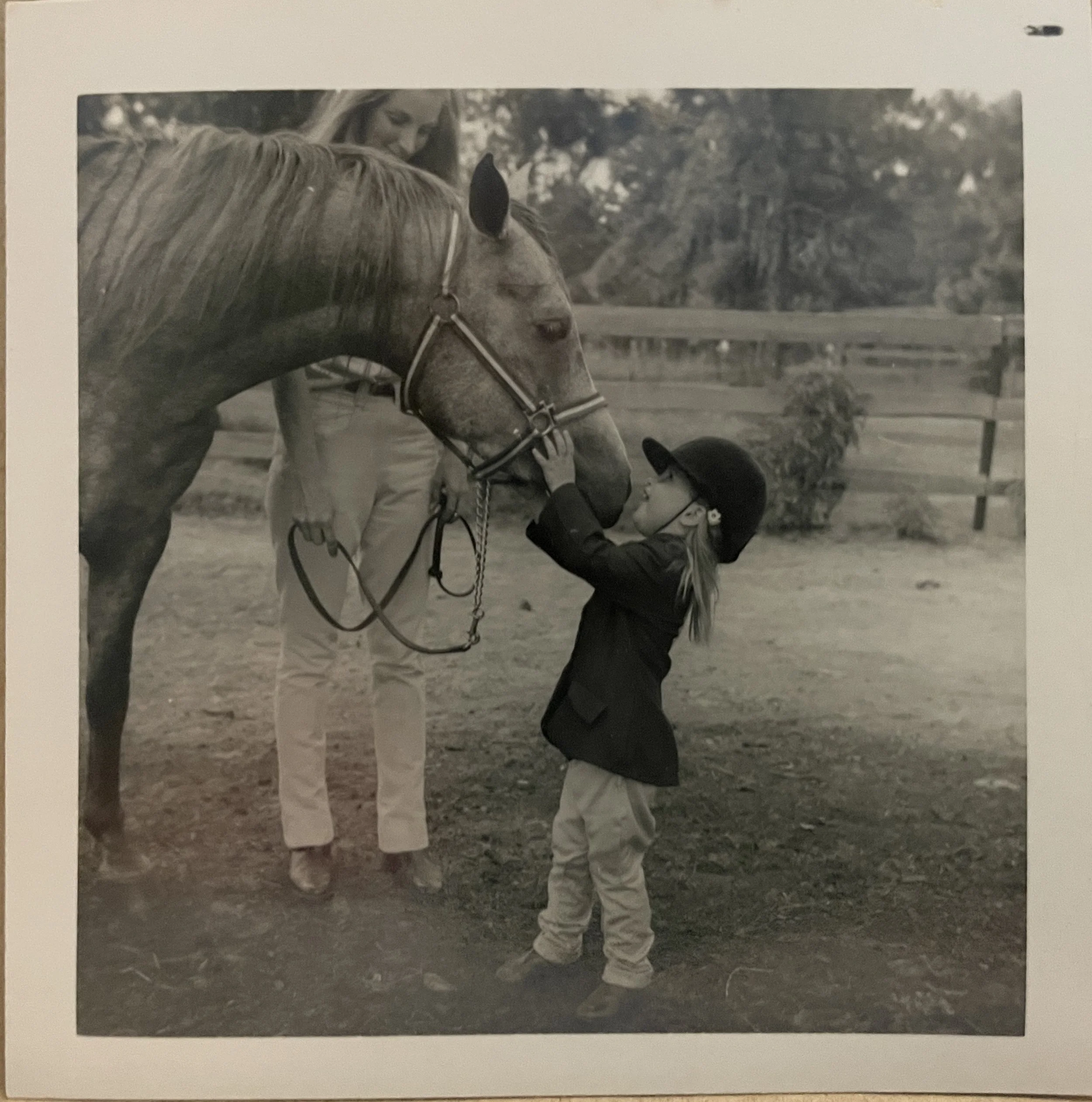 A person holding a horse's bridle near a child in an outdoor setting with trees and a wooden fence in the background.