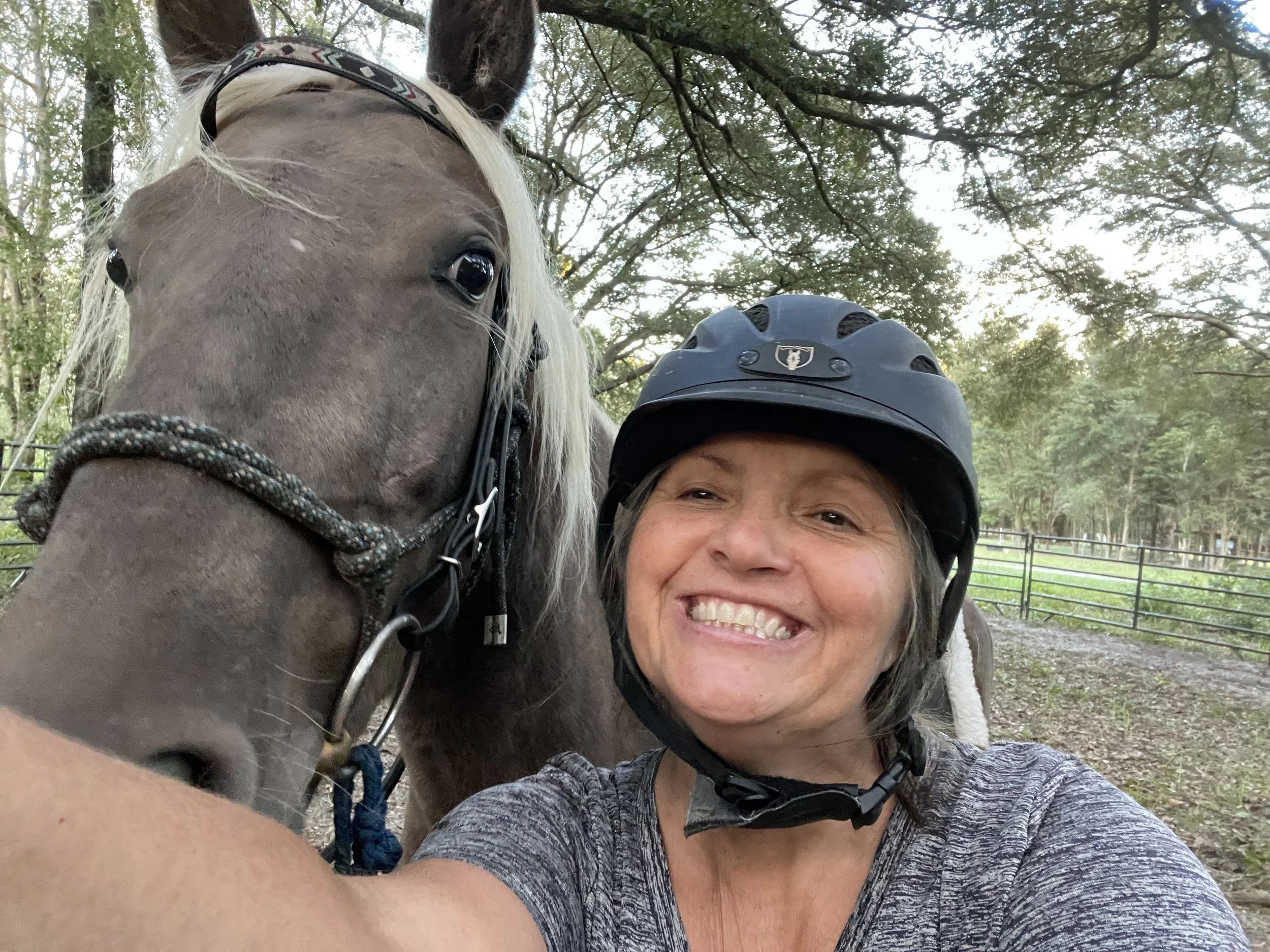 A woman smiling and taking a selfie with a horse at an outdoor location. The horse's face is close to the camera, and the woman is wearing a helmet. There are trees and a fence in the background.
