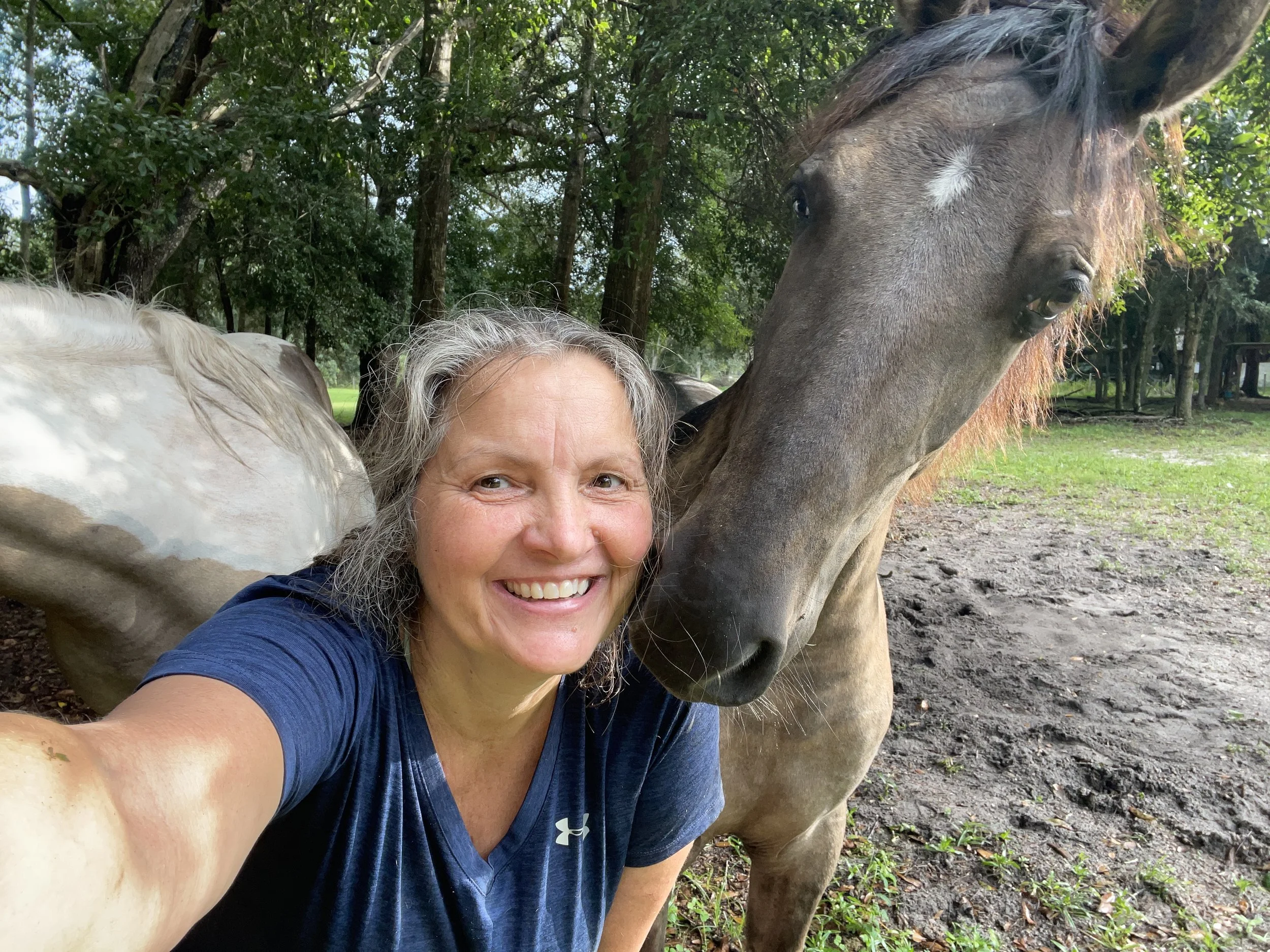 A woman with curly gray hair taking a selfie with a gray horse in a wooded area.