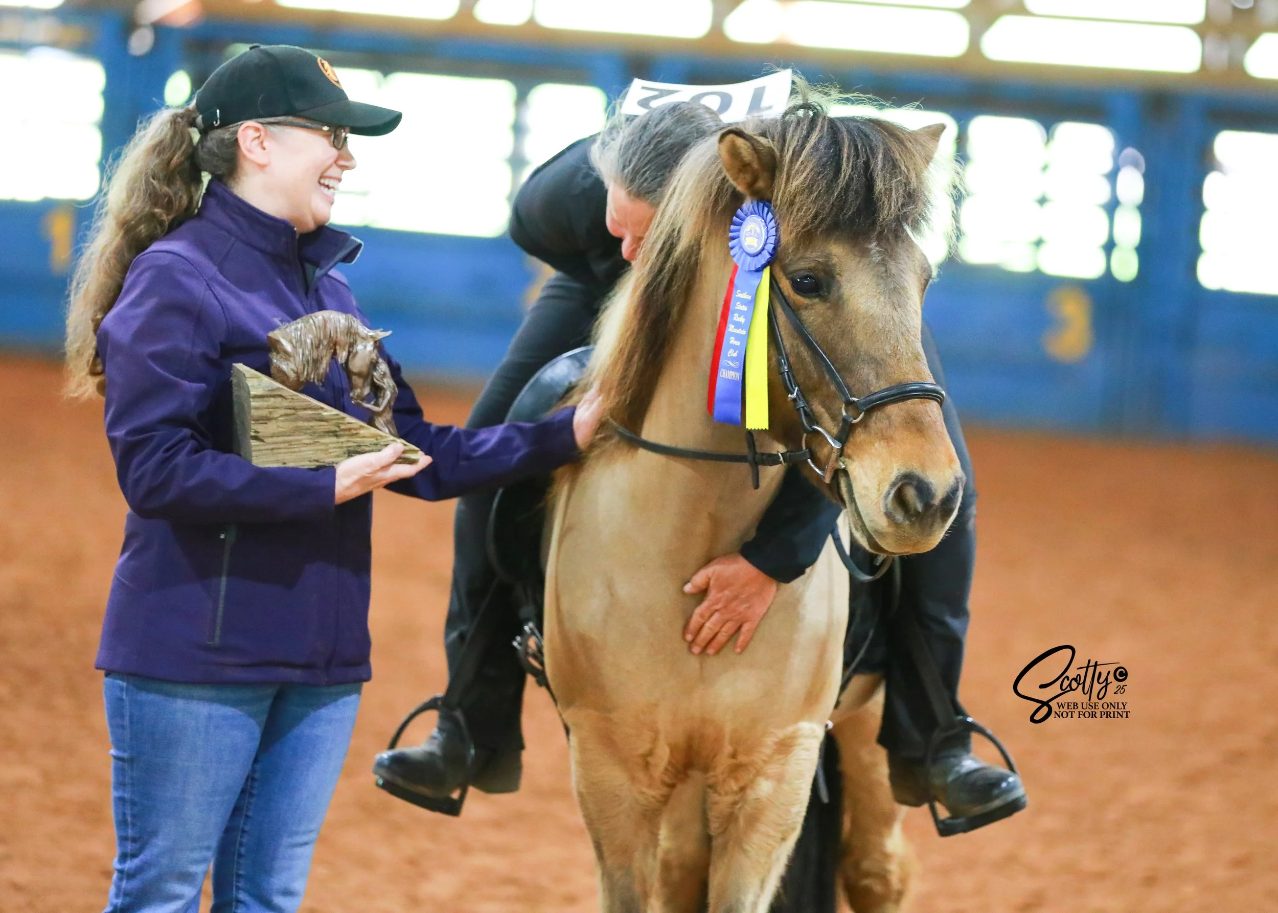 Woman holding a trophy of a horse with a person in a black jacket leaning on the horse's neck at an indoor equestrian event.