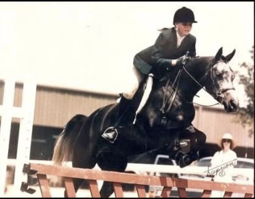Young woman riding a dark horse jumping over an obstacle during an equestrian event, with person in sunglasses and hat standing nearby.