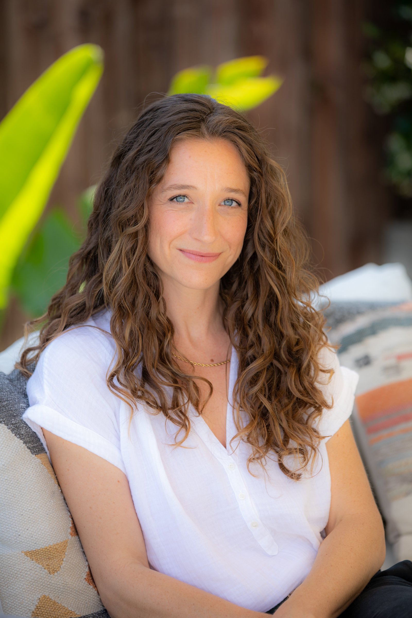Julie Marx, a woman with long, curly brown hair and blue eyes, wearing a white shirt and gold necklace, sitting outdoors with a blurred background of greenery.