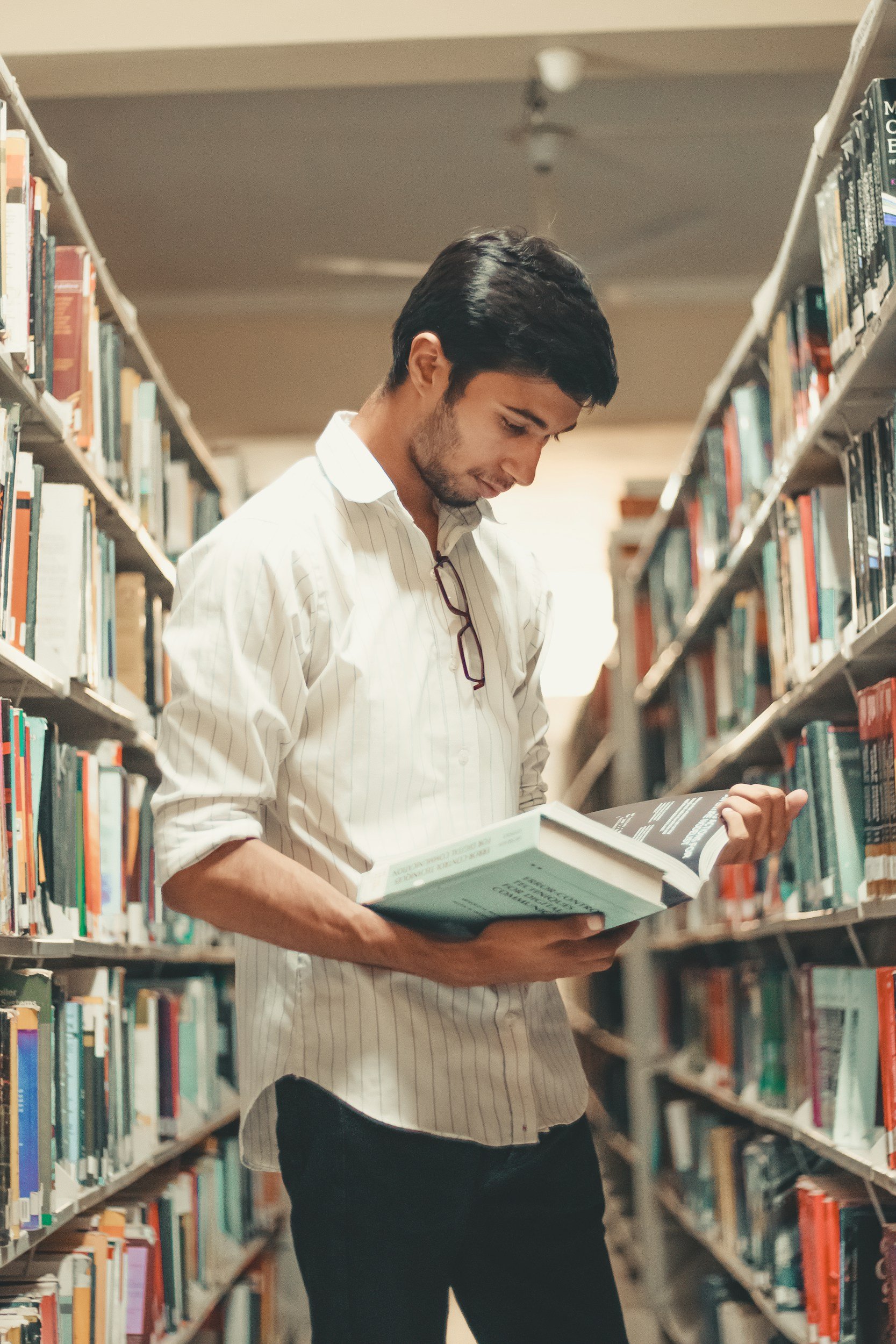 A young man with dark hair, a beard, and a white striped shirt stands in a library aisle, looking down at a book he is holding.