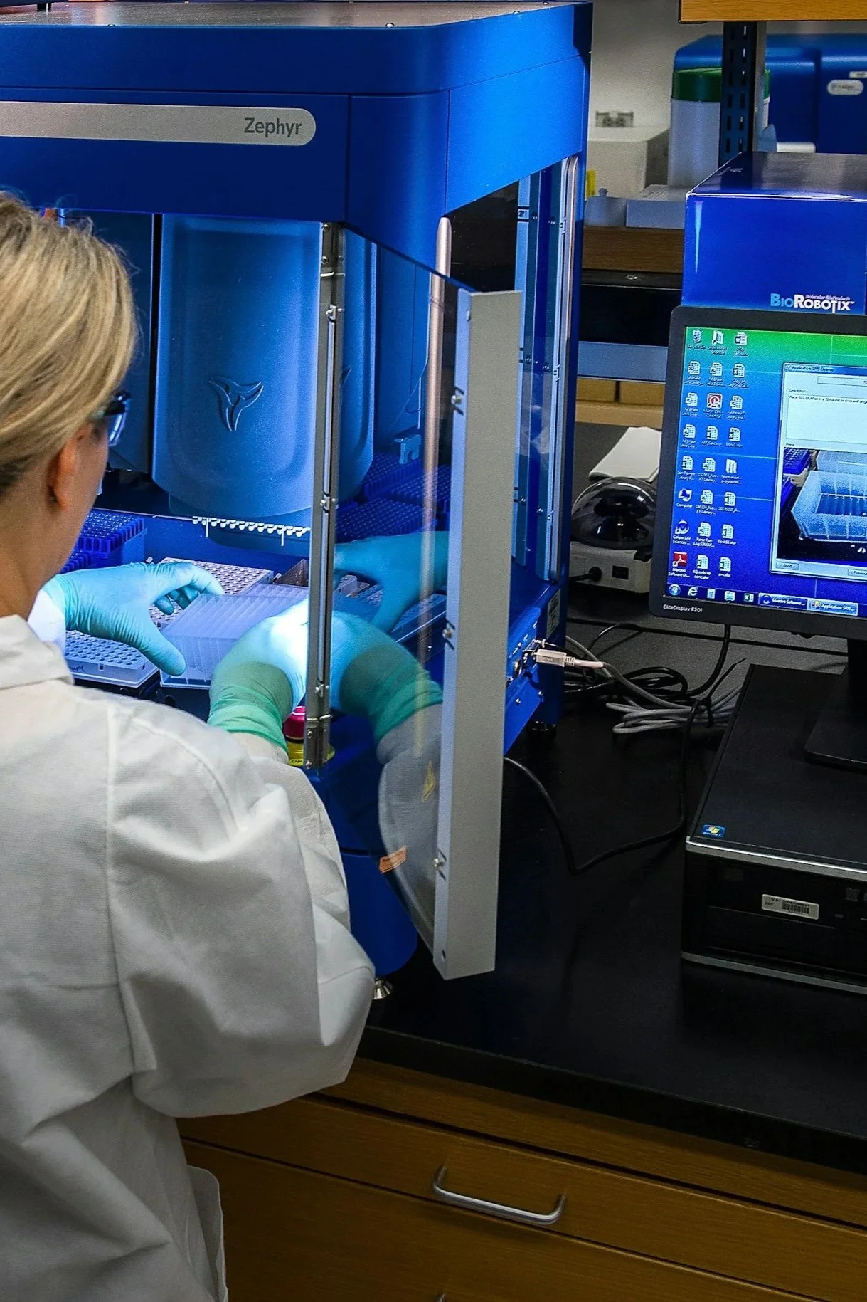 A scientist working with a laboratory machine labeled 'ZephyR' and a computer monitor displaying data and graphs in a laboratory setting.