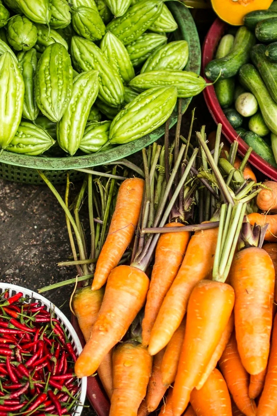 Fresh bitter melons, carrots, cucumbers, and red chili peppers displayed at a market.